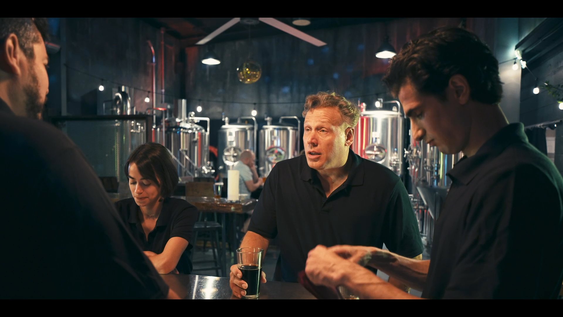 Brewery staff members discuss operations and review product samples while gathered around a service counter. The team engages in a professional interaction focused on training and quality assessment in a production environment.