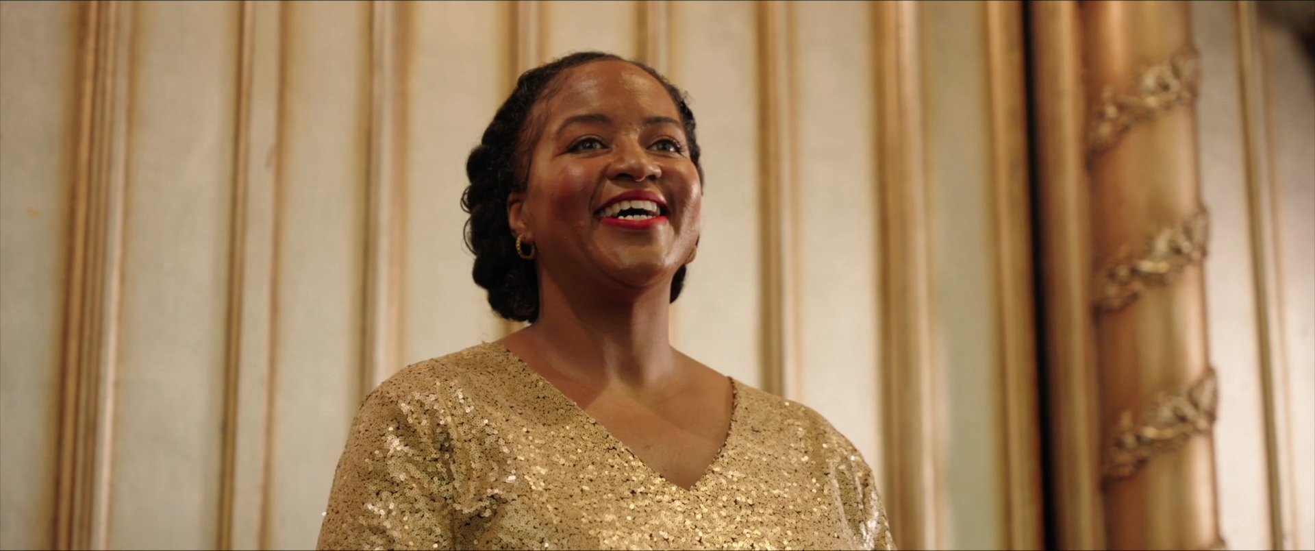 A singer wearing a gold sequin dress performs an expressive musical piece before a gathered audience in a church. The congregation responds with enthusiastic applause, highlighting a collaborative and celebratory community atmosphere.