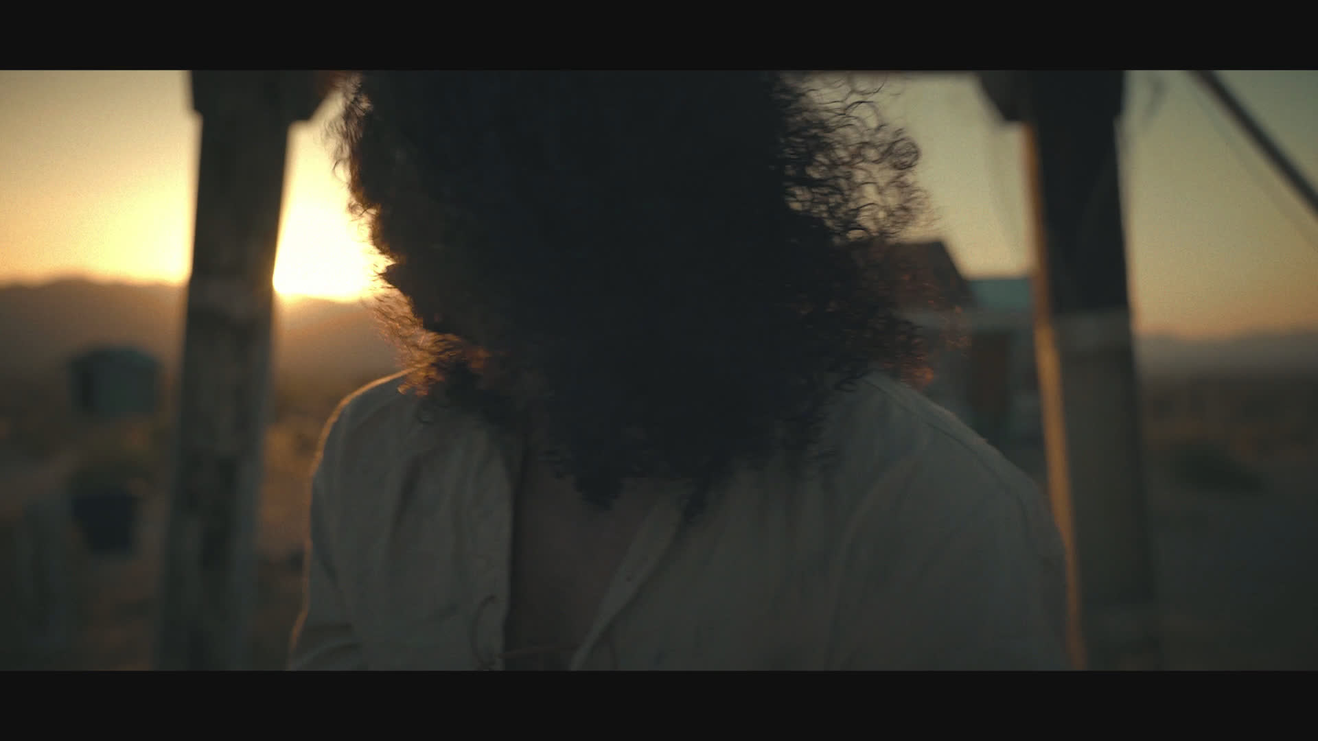 At sunset in a dusty frontier settlement, a woman in a black cowboy hat raises and fires a revolver as a curly-haired figure reacts nearby. She lowers the gun and speaks with dry composure amid the tense confrontation.