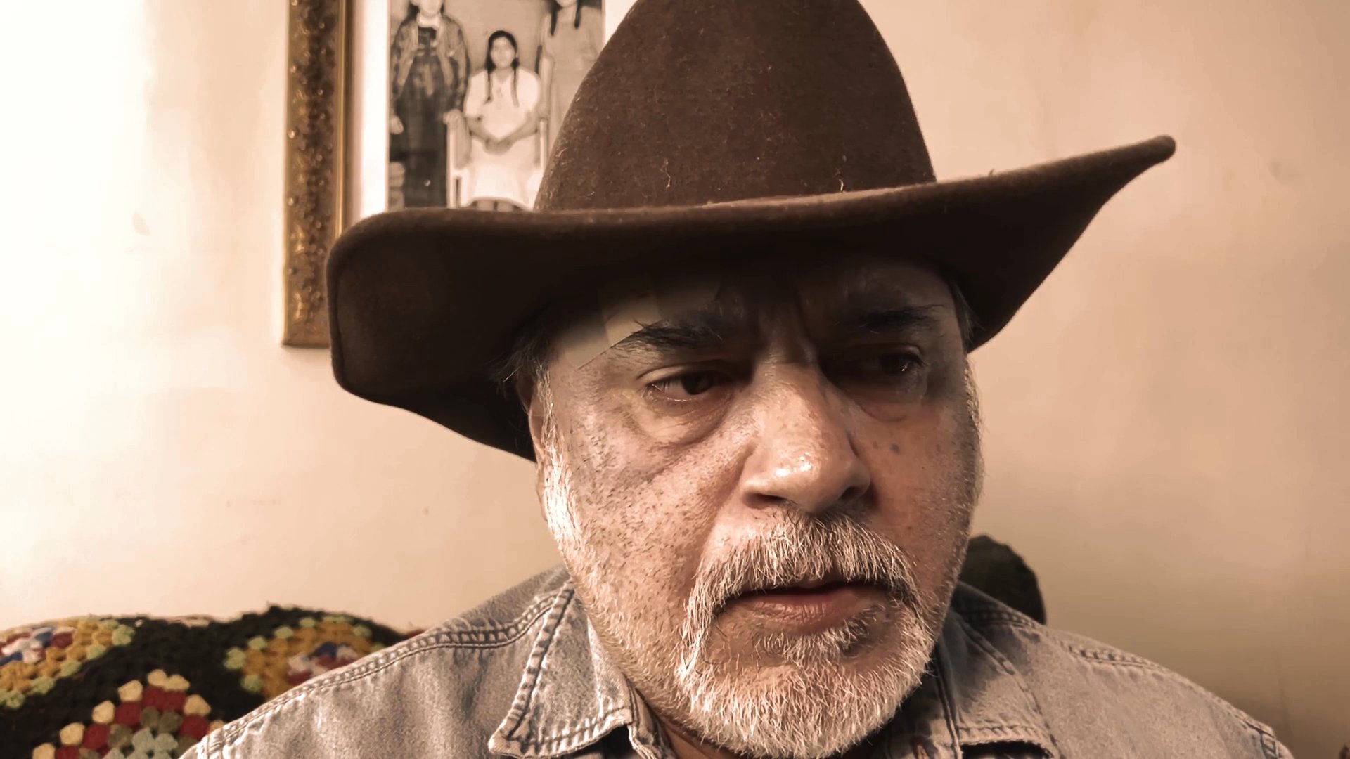 A man wearing a cowboy hat delivers a direct, conversational monologue while seated indoors against a domestic backdrop.