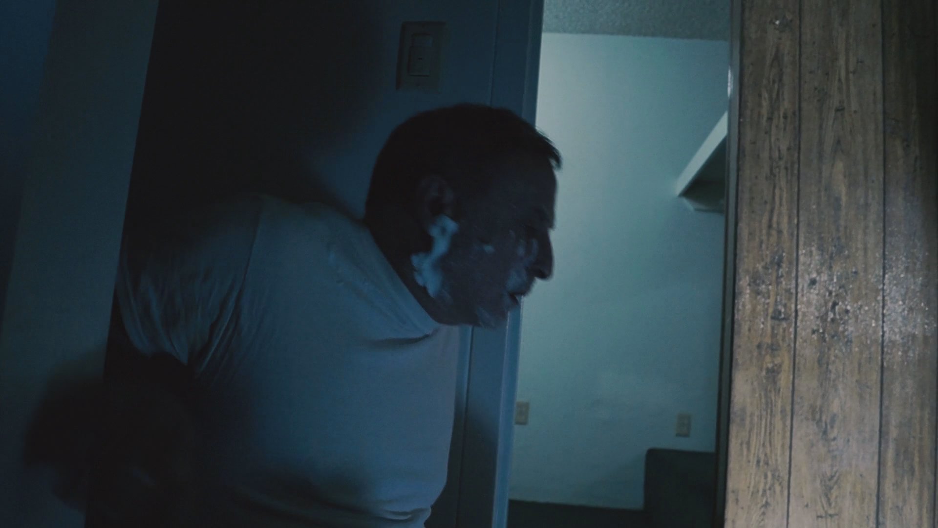 A man applying shaving cream to his face in a dimly lit bathroom suddenly becomes startled and abruptly leaves the room. His routine is interrupted by an urgent development, prompting him to move quickly away from the mirror.