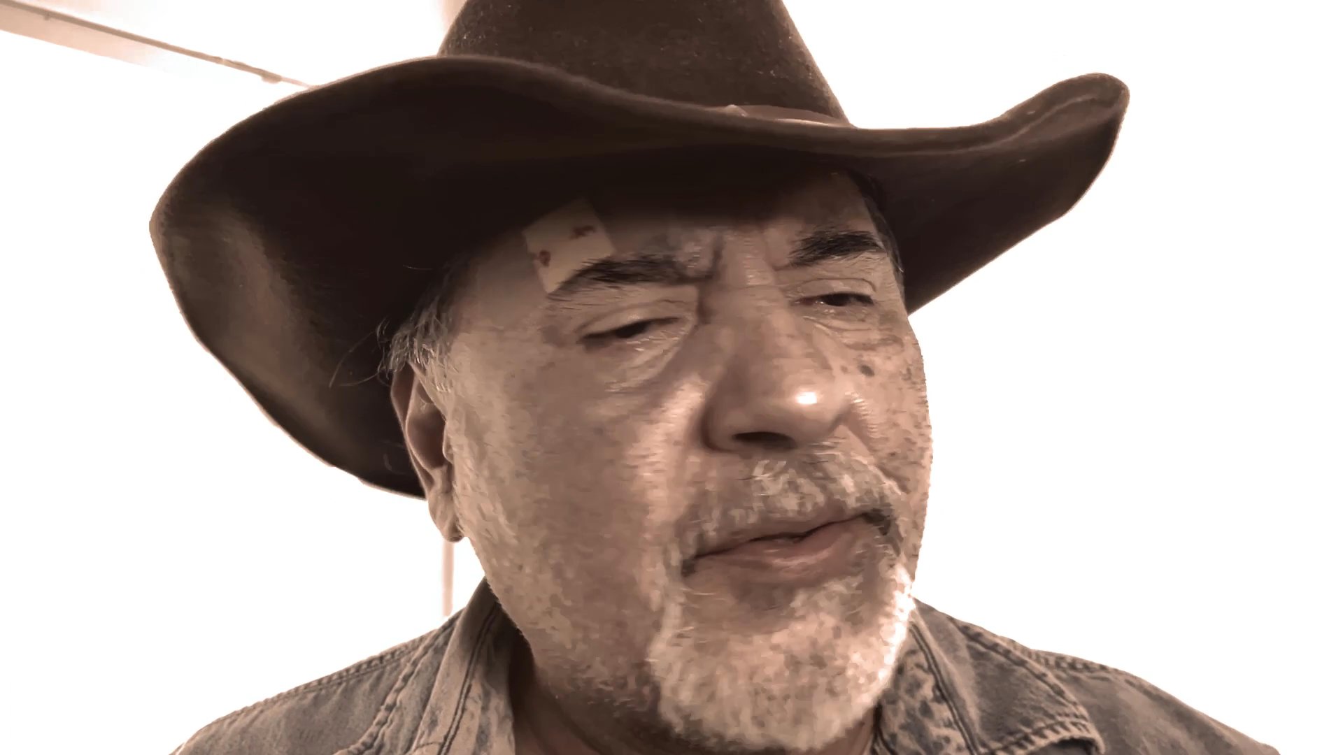 A man wearing a cowboy hat speaks directly to the camera in a close-up, expressive monologue.