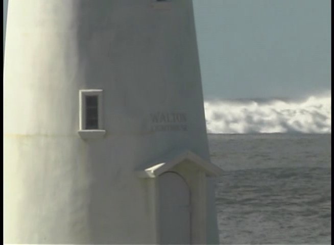 A surfer rides waves near a historic lighthouse while a tall ship navigates the foggy waters nearby.