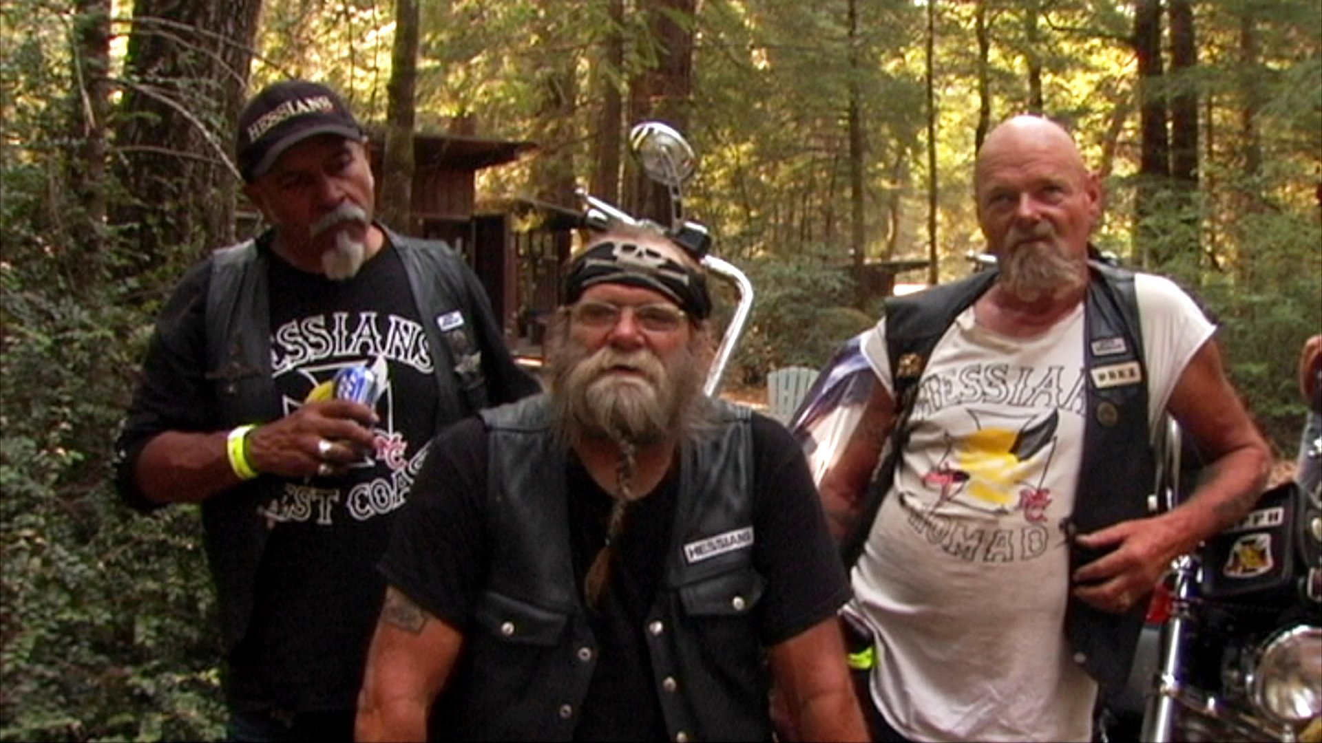Members of the Hessians motorcycle club discuss their experiences and affiliations in an outdoor, wooded setting. The segment features individual portraits and close-ups of the men wearing signature club vests and patches.