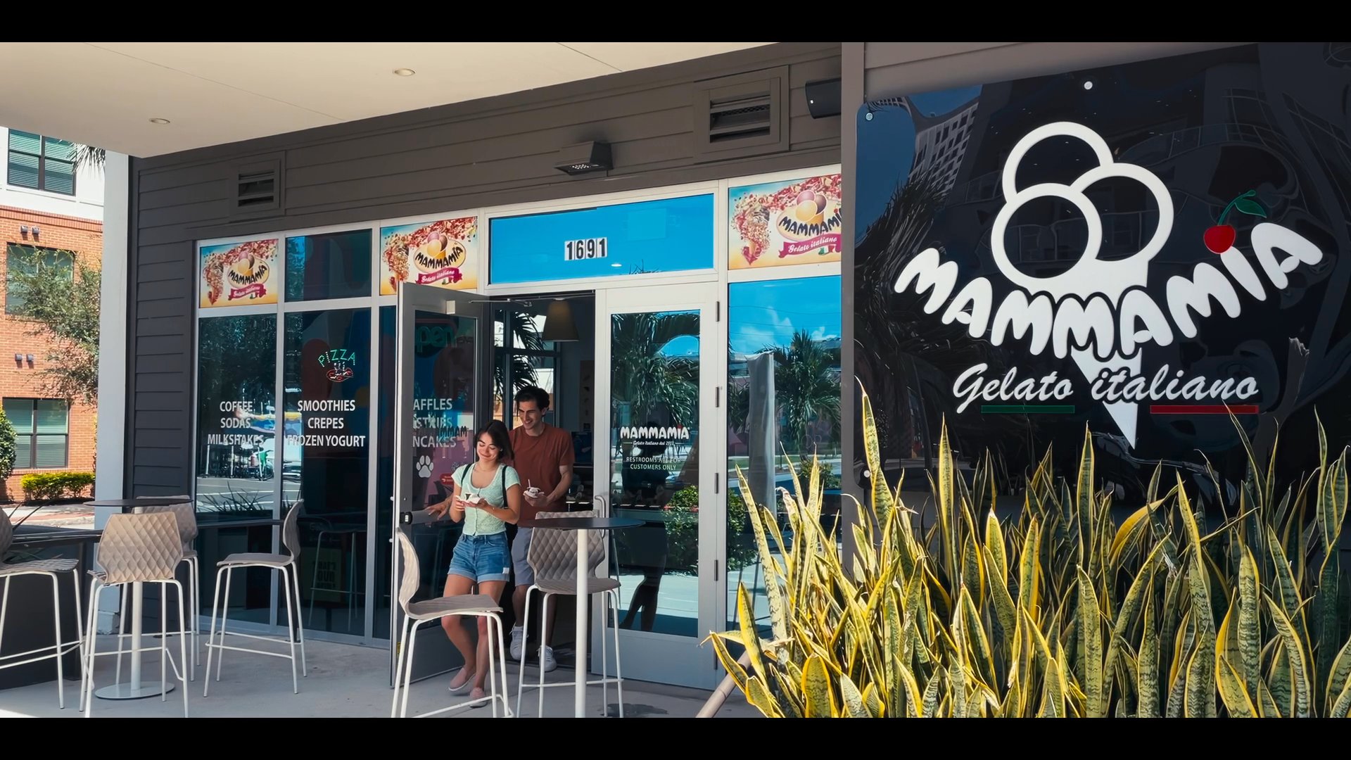 A couple enjoys gelato at an outdoor table in front of the Mammamia Gelato Italiano shop in St. Pete. The scene highlights the storefront atmosphere and the relaxed dining experience.