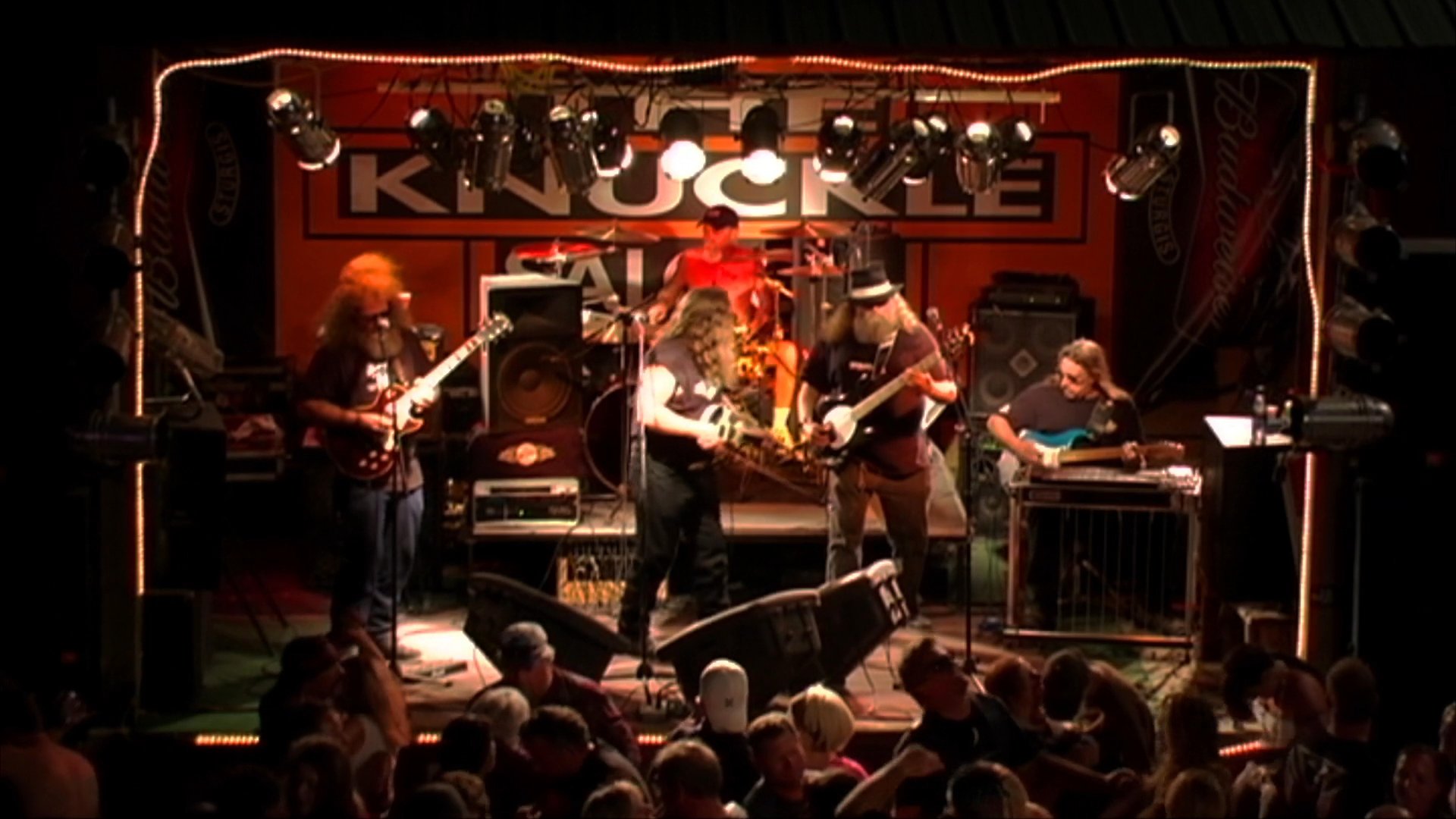 A rock band performs on a stage at a bar while a crowd dances and socializes in the venue. The footage captures the high-energy atmosphere of the musical event.