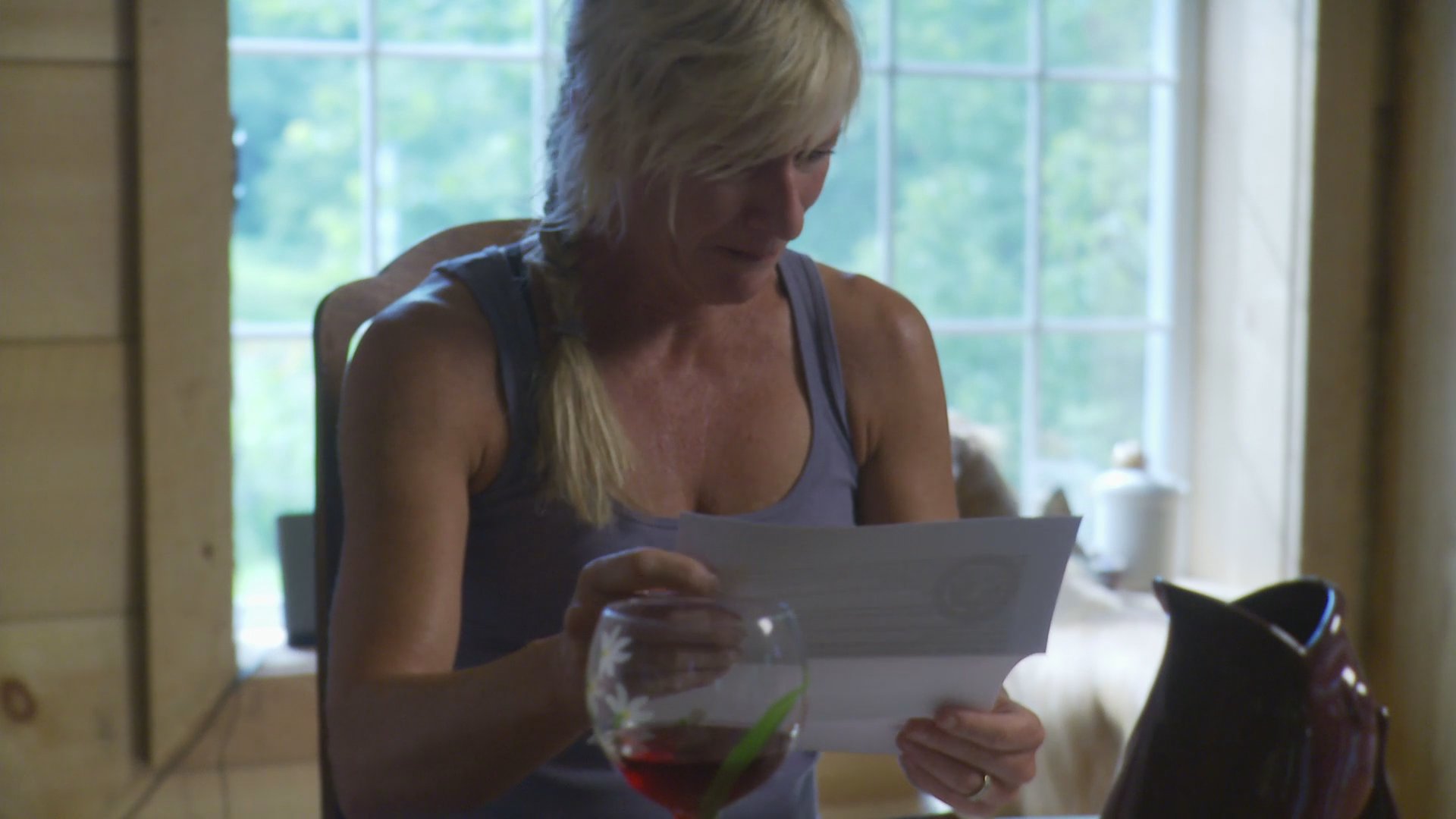 A woman sits by a window intently reading a document. She appears focused as she reviews the pages in a quiet indoor setting.