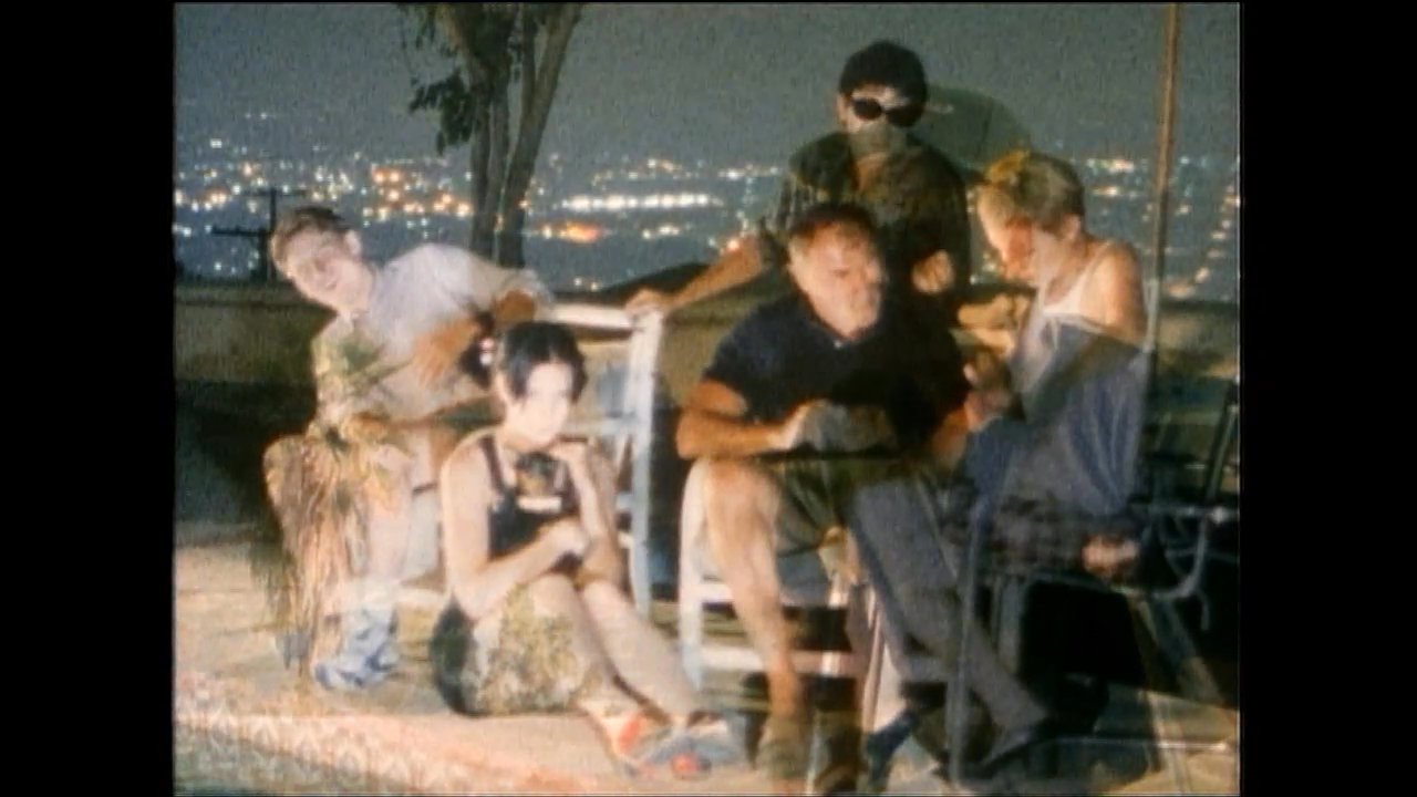 An injured man using crutches transitions from walking on a rooftop terrace at dusk to sitting and conversing with a group of friends as the city lights appear.