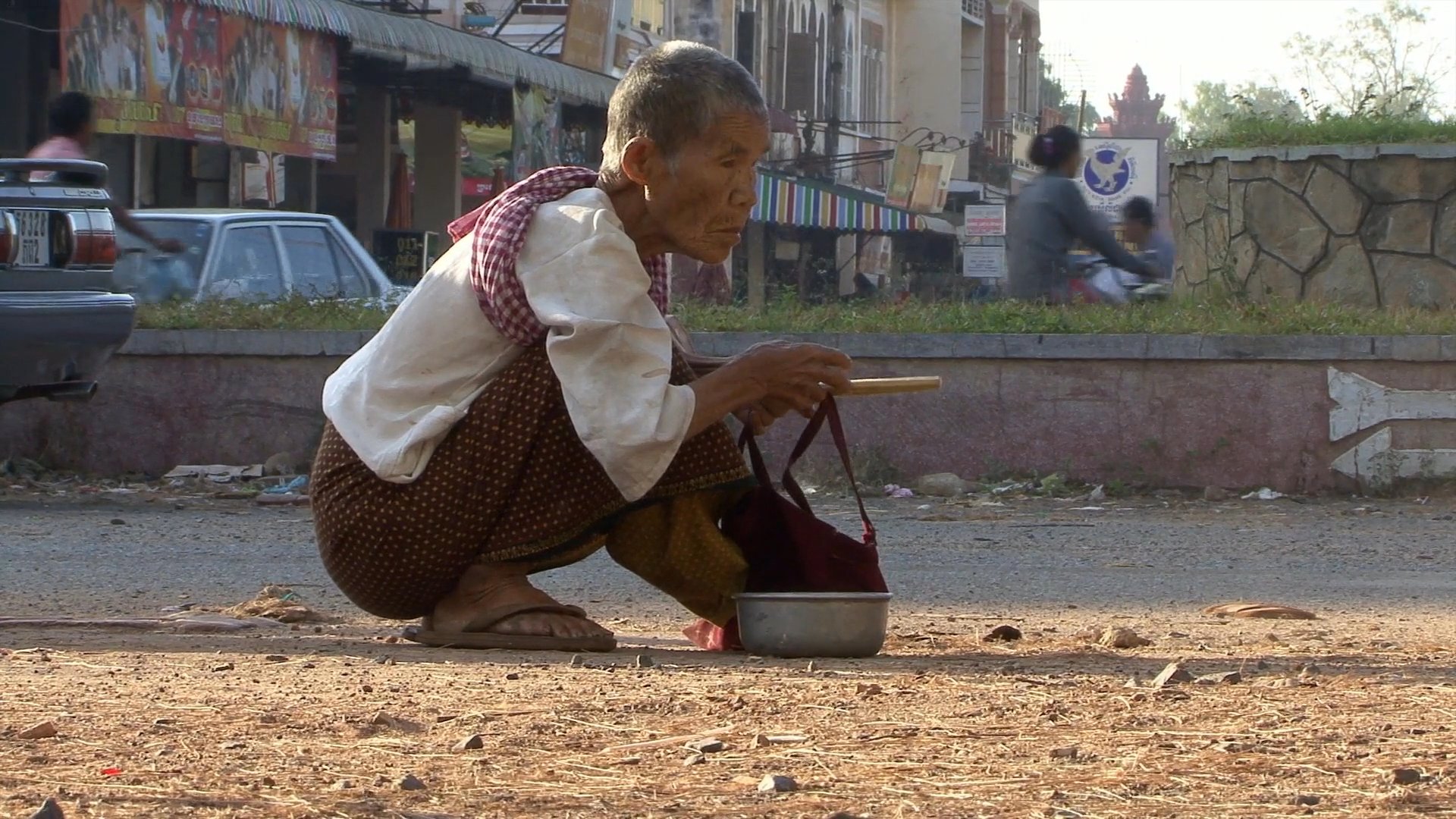 An elderly person sits on a roadside in a busy urban area counting currency while traffic passes by, contrasted with a nearby child playing in the street.