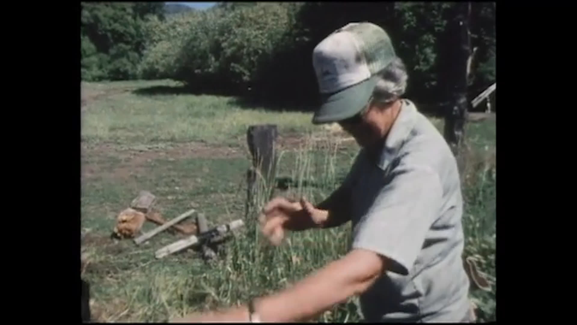 In a grassy clearing, a man gestures to demonstrate how a chainsaw kicked back and struck his face, describing the impact across his glasses and into his mouth and teeth. Cut wood lies nearby as another person listens.