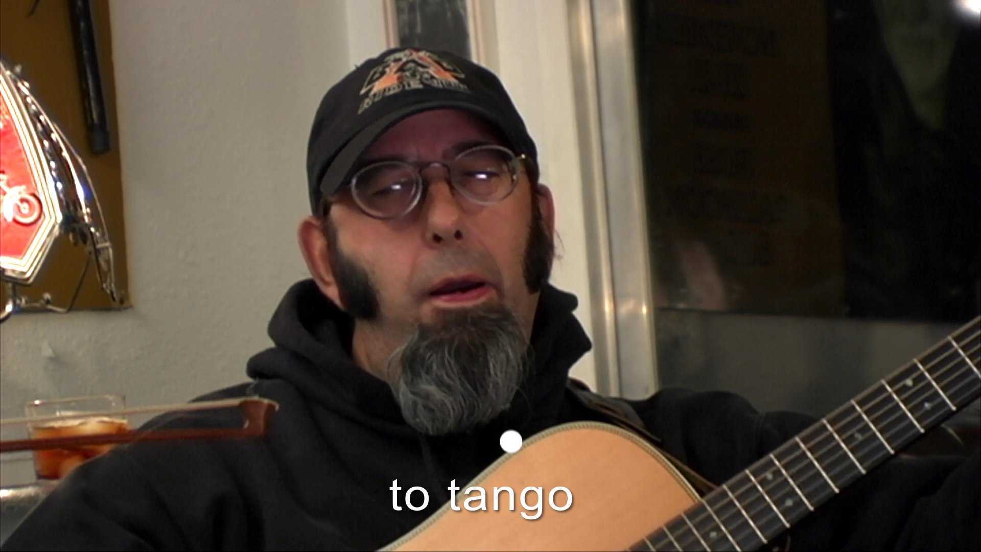 A man with a goatee and baseball cap sings and plays acoustic guitar, accompanied by another musician in a casual indoor setting.