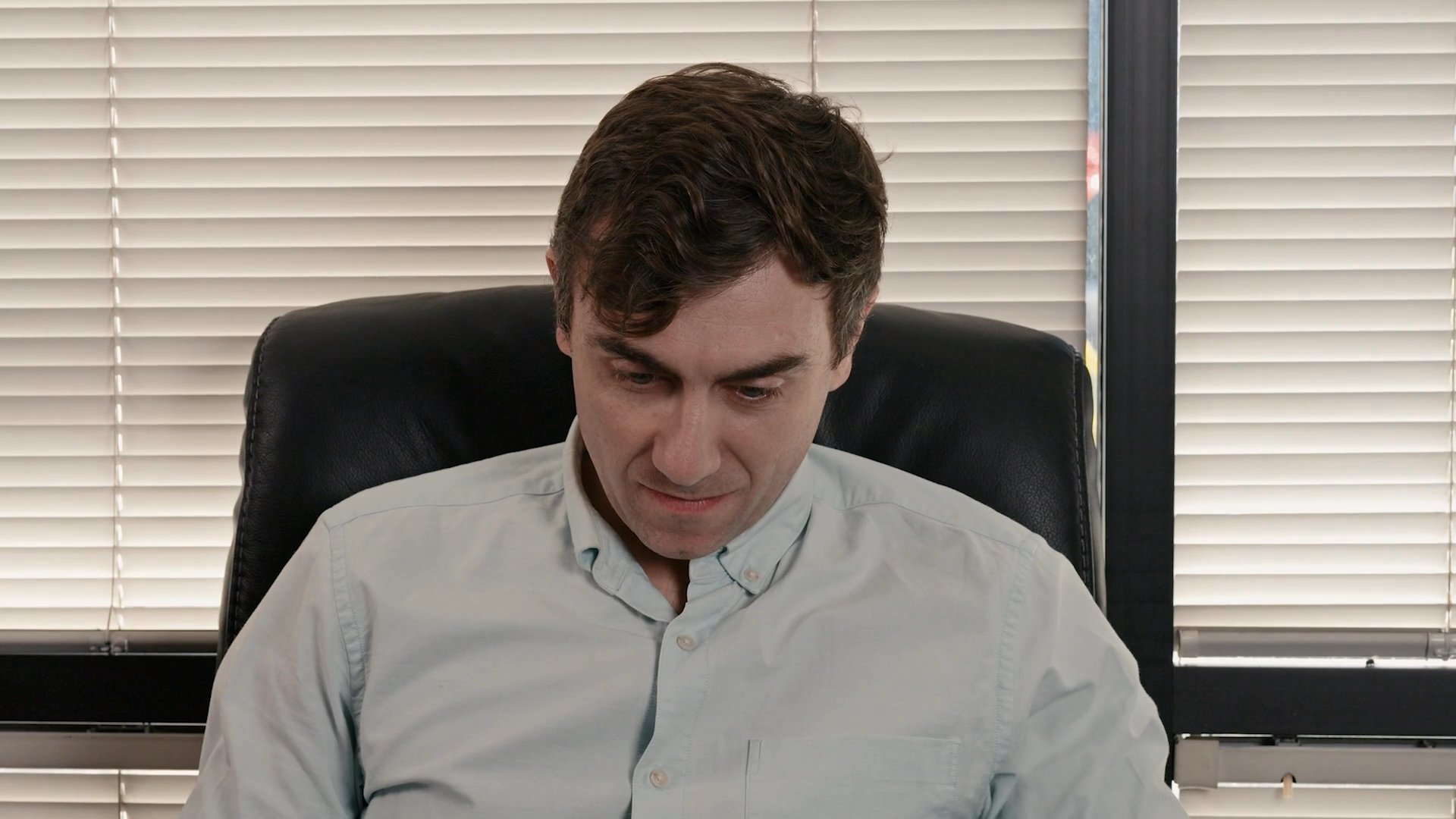 A man in a light-colored button-down shirt engages in a professional conversation within an office setting. He maintains eye contact and gestures while speaking to another individual across a desk.
