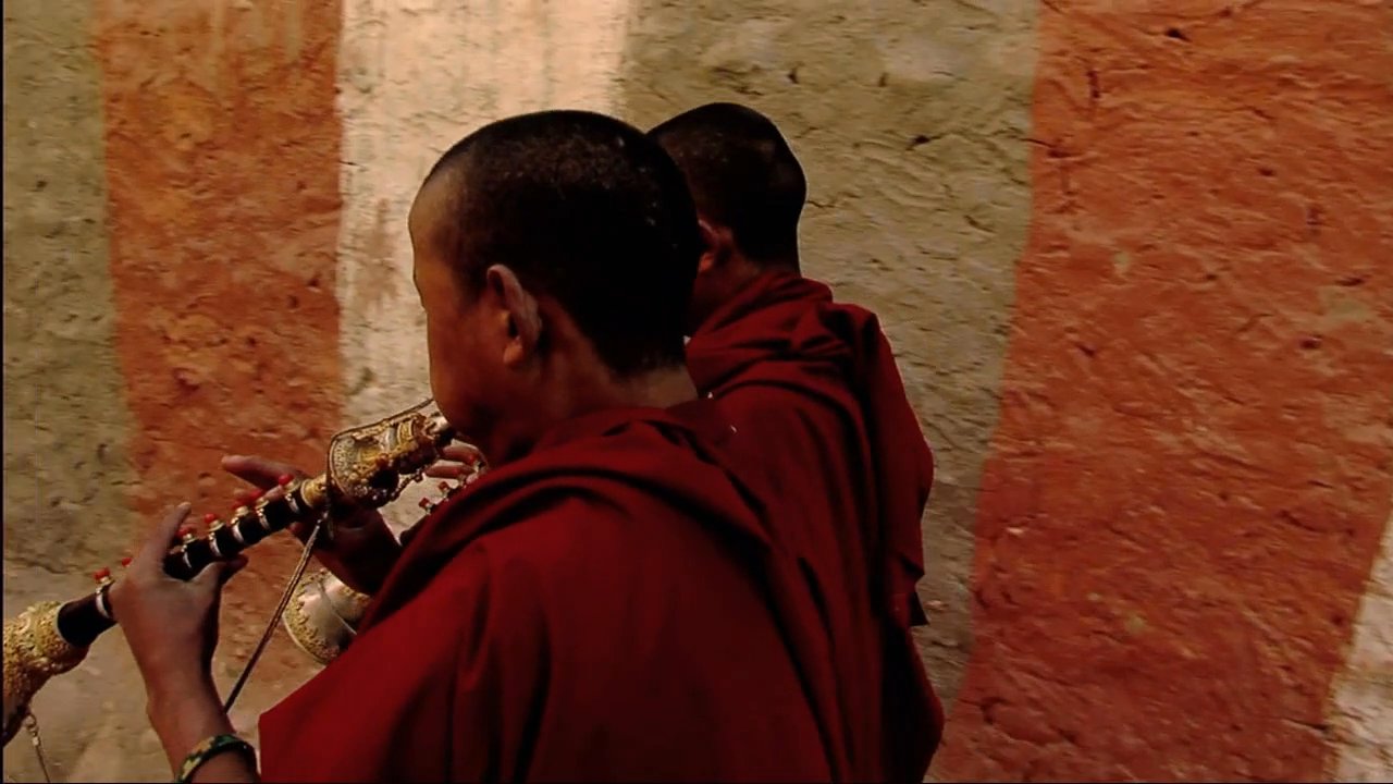Tibetan monks in traditional robes play ceremonial gyaling instruments within the grounds of a mountain monastery. The sequence captures their focused performance amidst the architectural setting, concluding with a serene reflection of the monastery in a nearby pond.