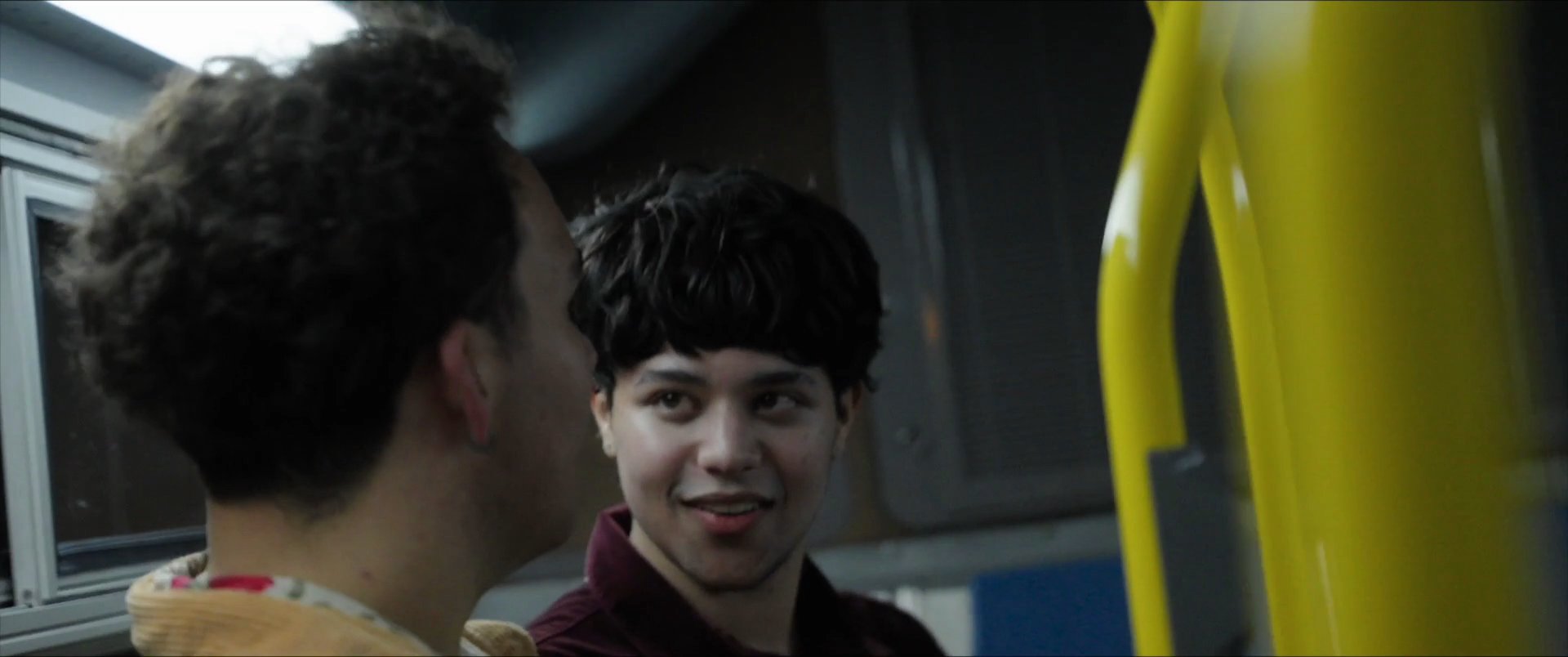 Two young men engage in an intimate conversation while sitting together on a public bus. Their focused eye contact and expressive facial reactions suggest a close personal connection.