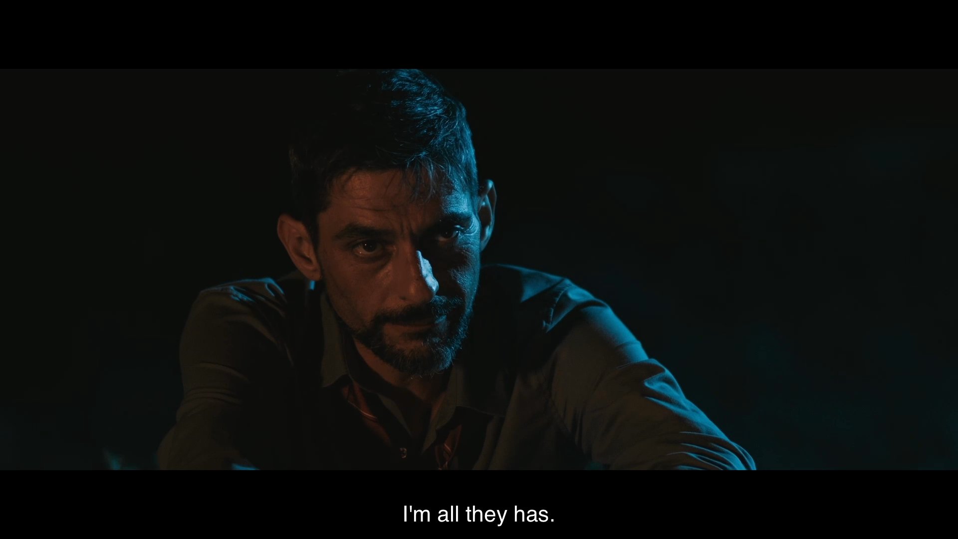 Two men sit by a campfire in the darkness, discussing personal loss and family dynamics in a somber, quiet exchange.