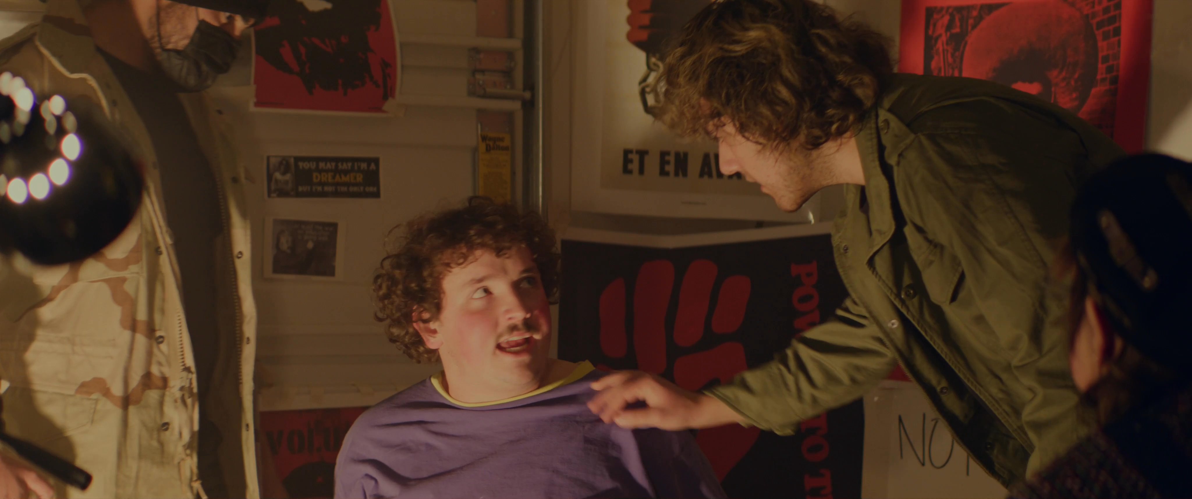 Young individuals engage in a dramatic, politically-charged roleplay within a room adorned with radical posters. The scene emphasizes intense, confrontational exchanges and themes of rebellion.