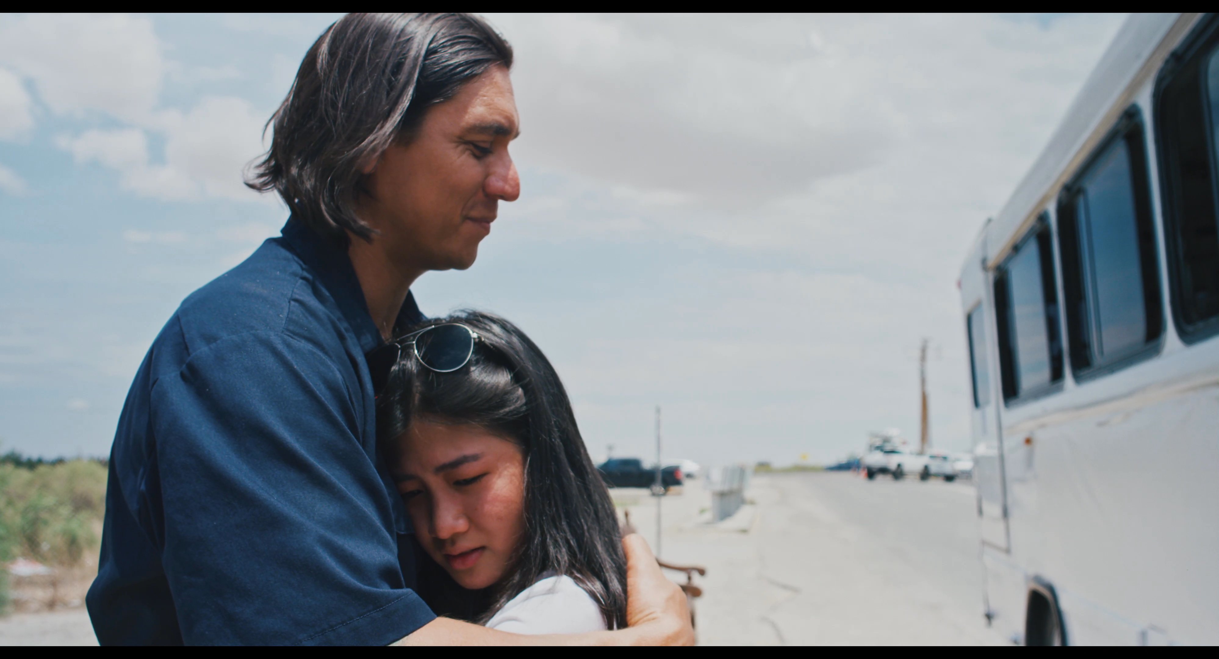 A man and a woman share an emotional embrace before the woman boards a waiting bus for her departure. The scene captures a poignant moment of separation in a roadside setting.