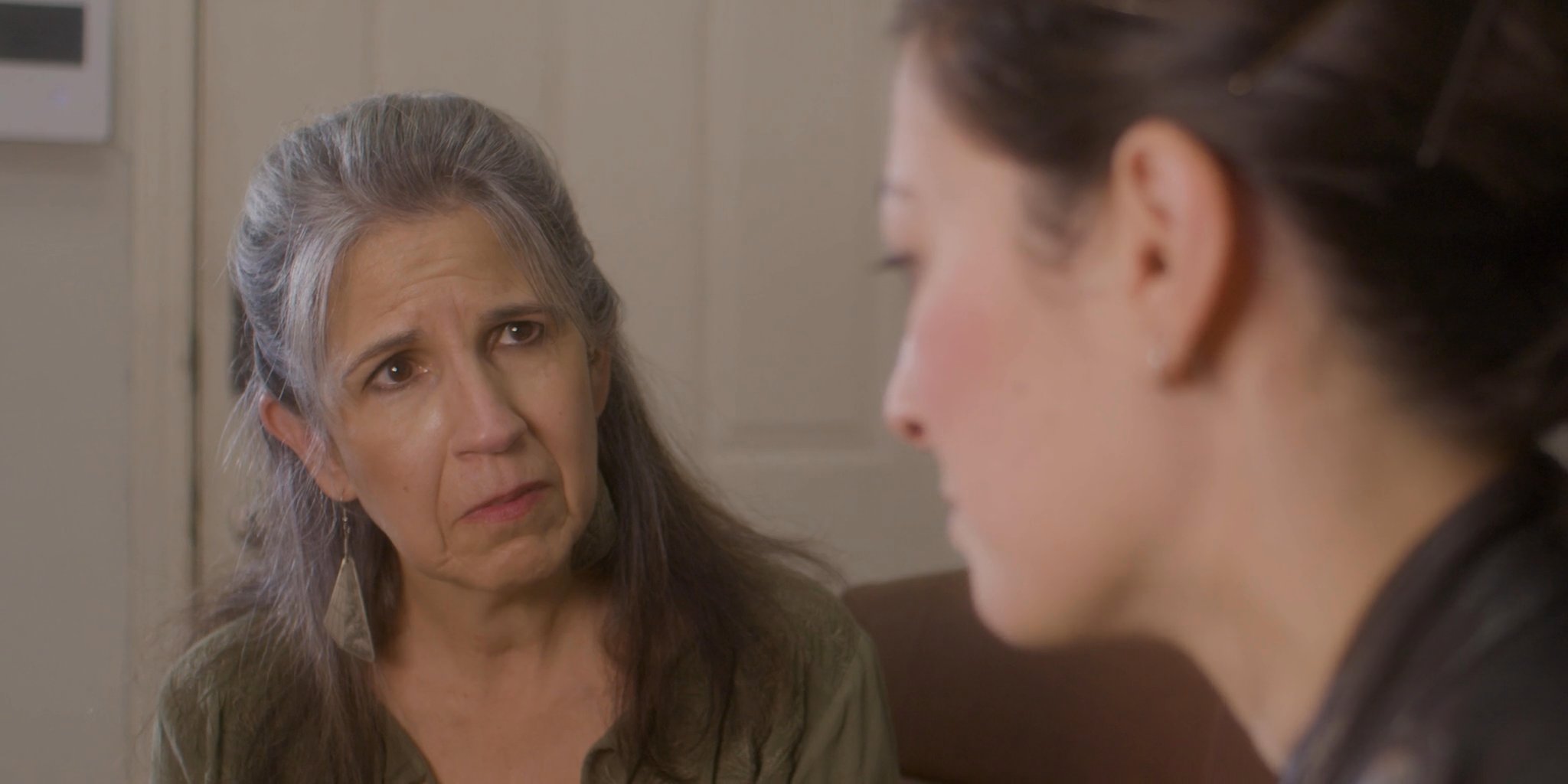 An older woman and a younger woman engage in an intimate, serious conversation within a home setting. The exchange reflects a significant emotional discussion between family members.