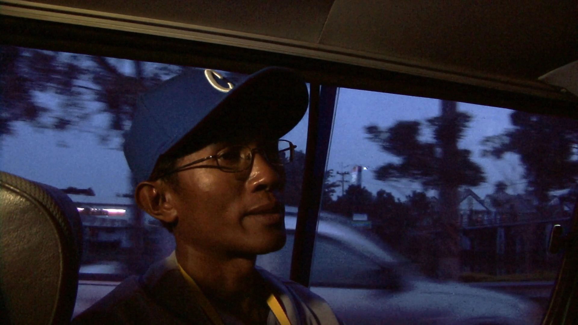 A man wearing a blue cap rides in the backseat of a vehicle during twilight, looking out the window while engaged in conversation. The passing landscape is visible through the window as the vehicle travels along a road.