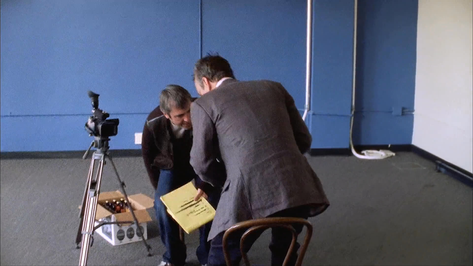 An actor adjusts a mascot headpiece while interacting with a director in a studio setting. The pair discusses the scene while preparing for a filming session.