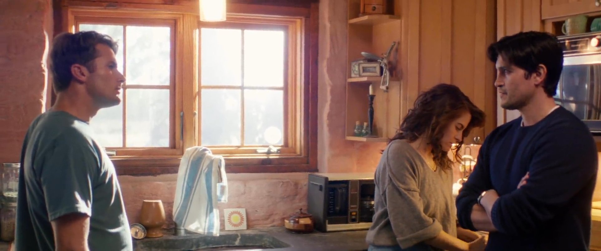 Three individuals engage in a tense, serious conversation within a domestic kitchen setting. The exchange involves focused dialogue and careful observation as they discuss an interpersonal situation.