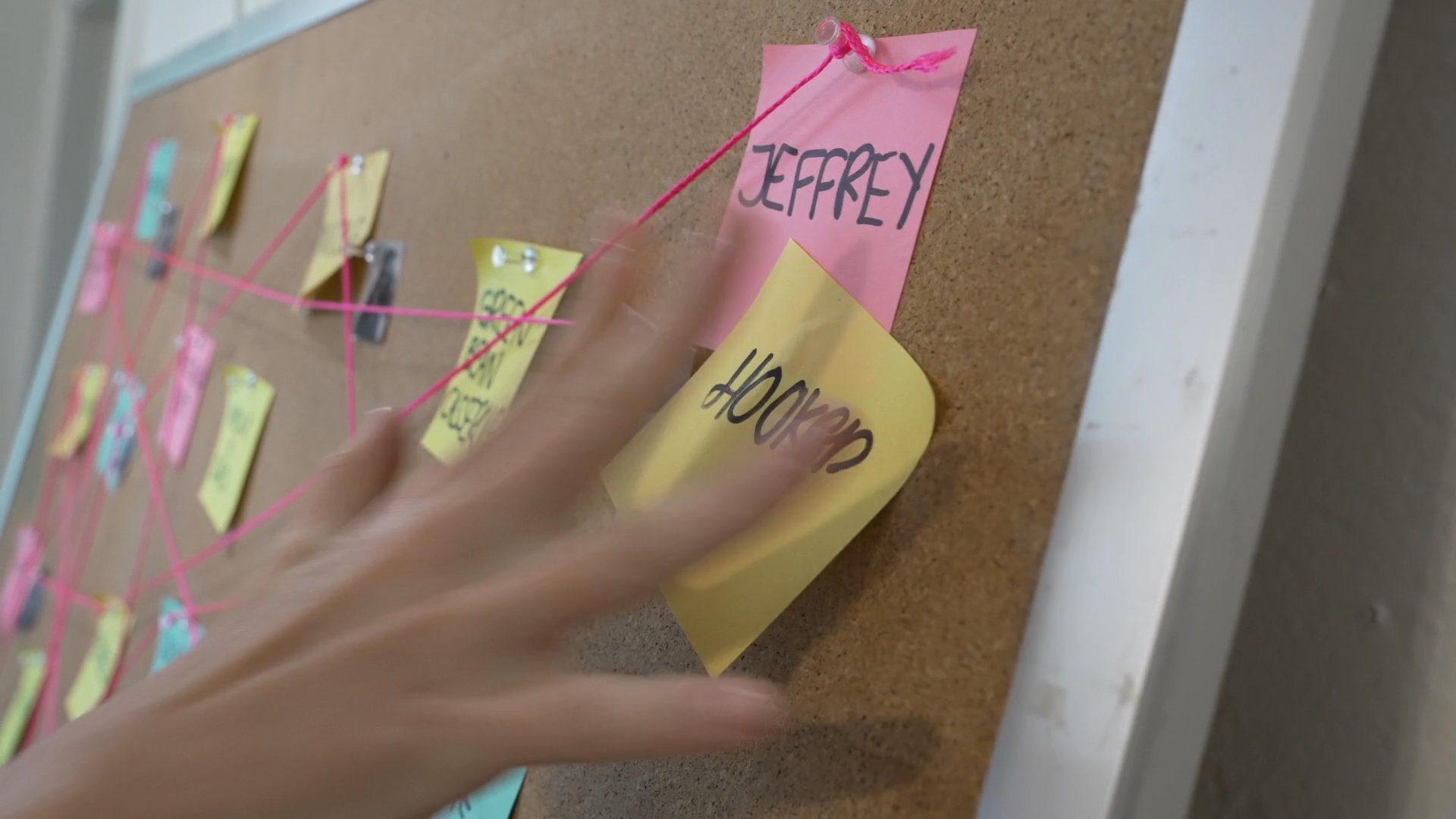 A woman meticulously organizes a complex web of connections on a corkboard as two men watch her from a nearby doorway. The scene transitions between outdoor activity and an intense, focused indoor investigation.