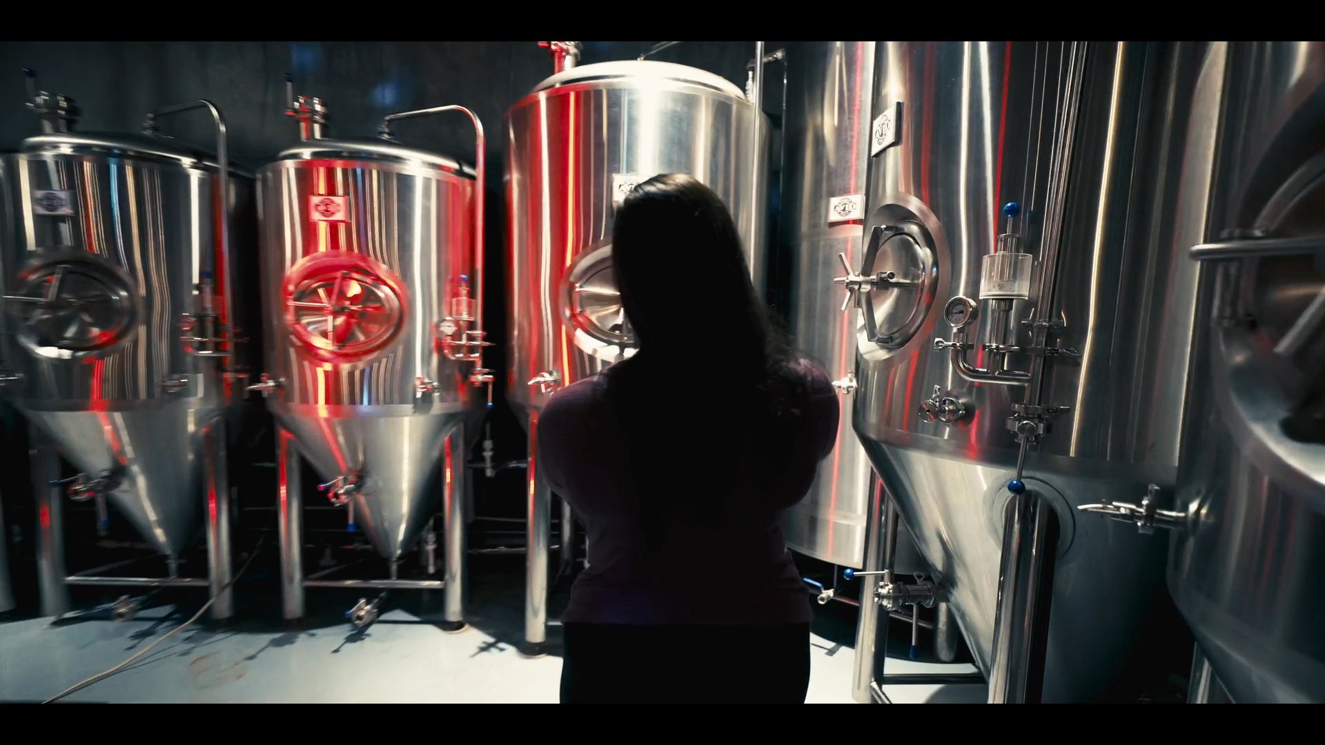 A technician conducts a routine inspection and sampling process within a commercial brewery, systematically checking stainless steel fermentation tanks.