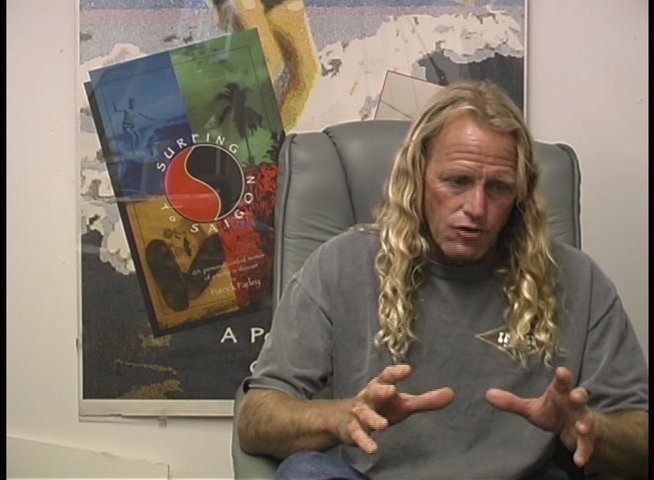 A man with long blonde hair discusses his experiences while sitting in front of a framed surfing poster. He utilizes expressive hand gestures to emphasize points during his commentary.