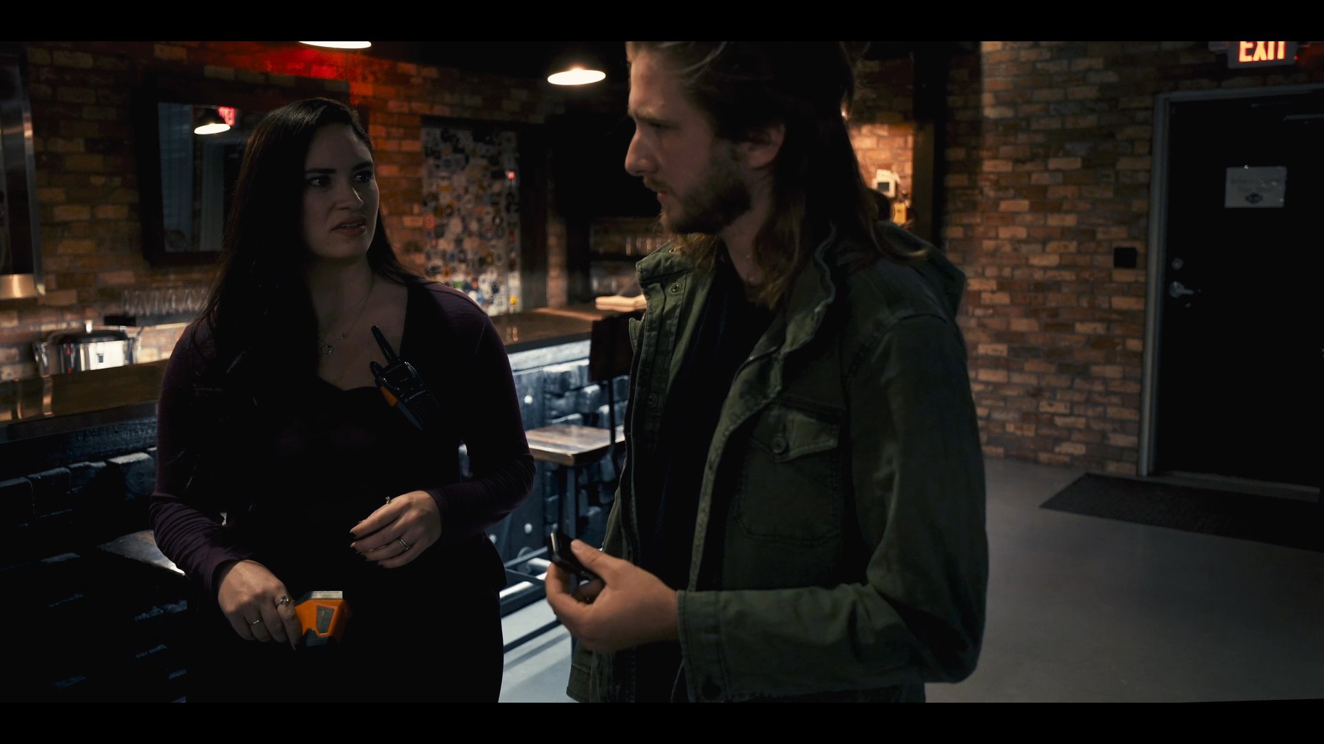 Two individuals engage in a professional discussion within an industrial-style interior featuring brick walls and ambient lighting. They exchange perspectives and gestures while maintaining an focused, collaborative interaction.