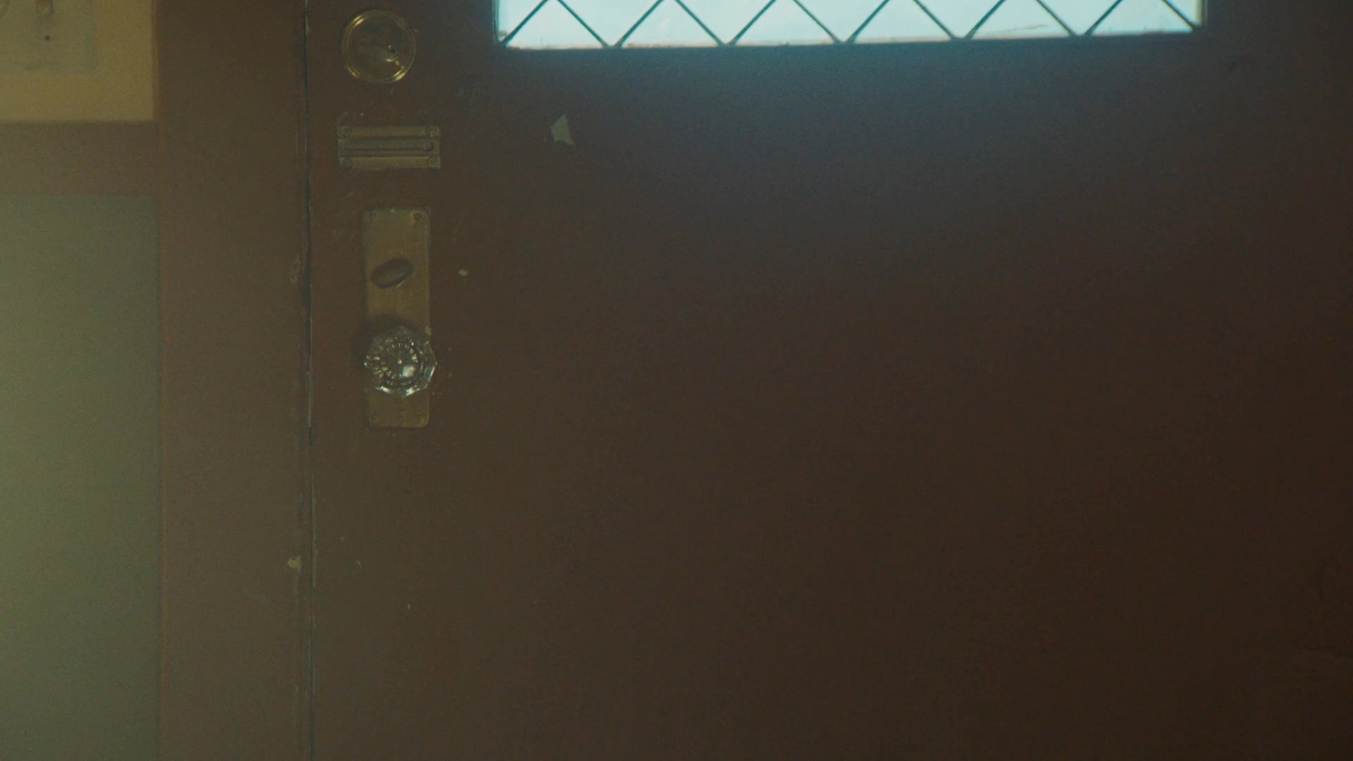 Two individuals enter a home through a front door and engage in an intimate, quiet conversation in the hallway. The setting suggests a private, domestic atmosphere as they arrive from outside.