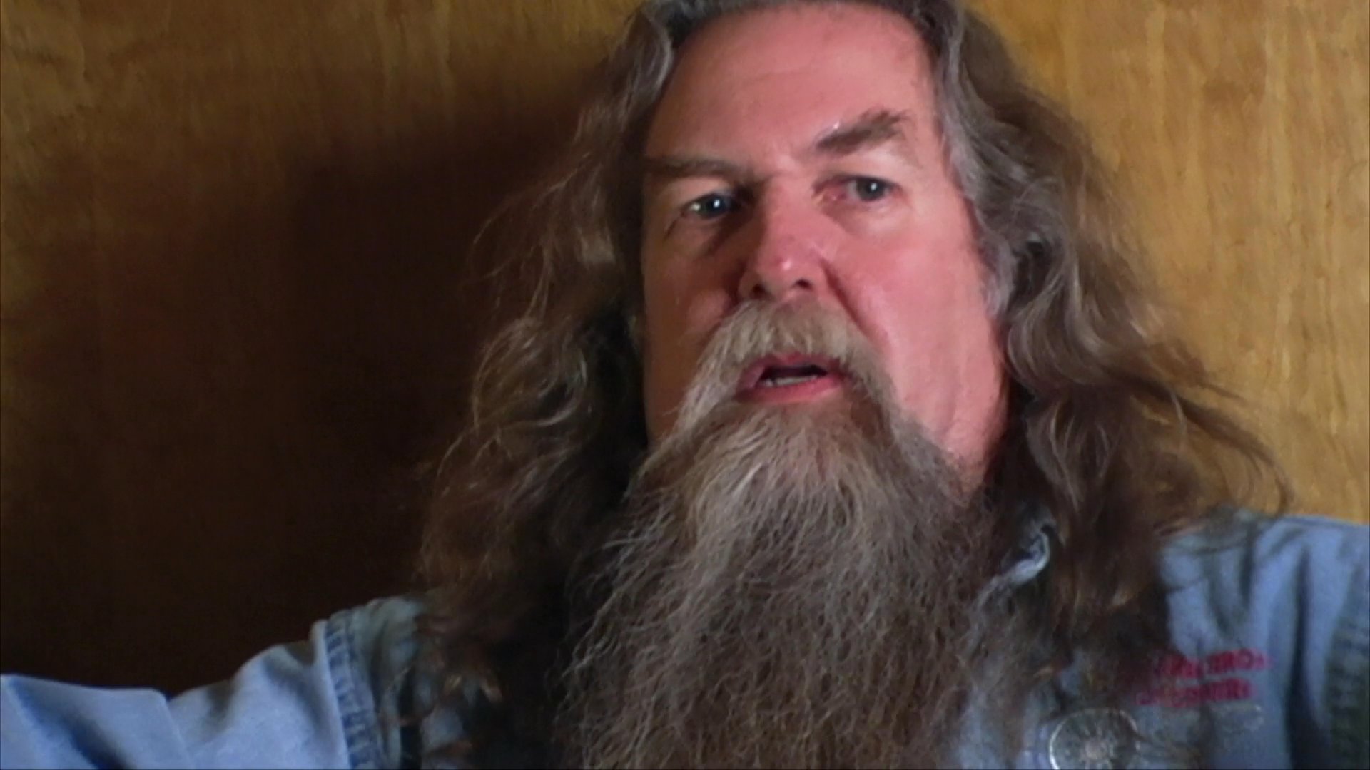 A man with a long beard recounts personal experiences and insights related to his work as a violin maker while sitting in a workshop setting.