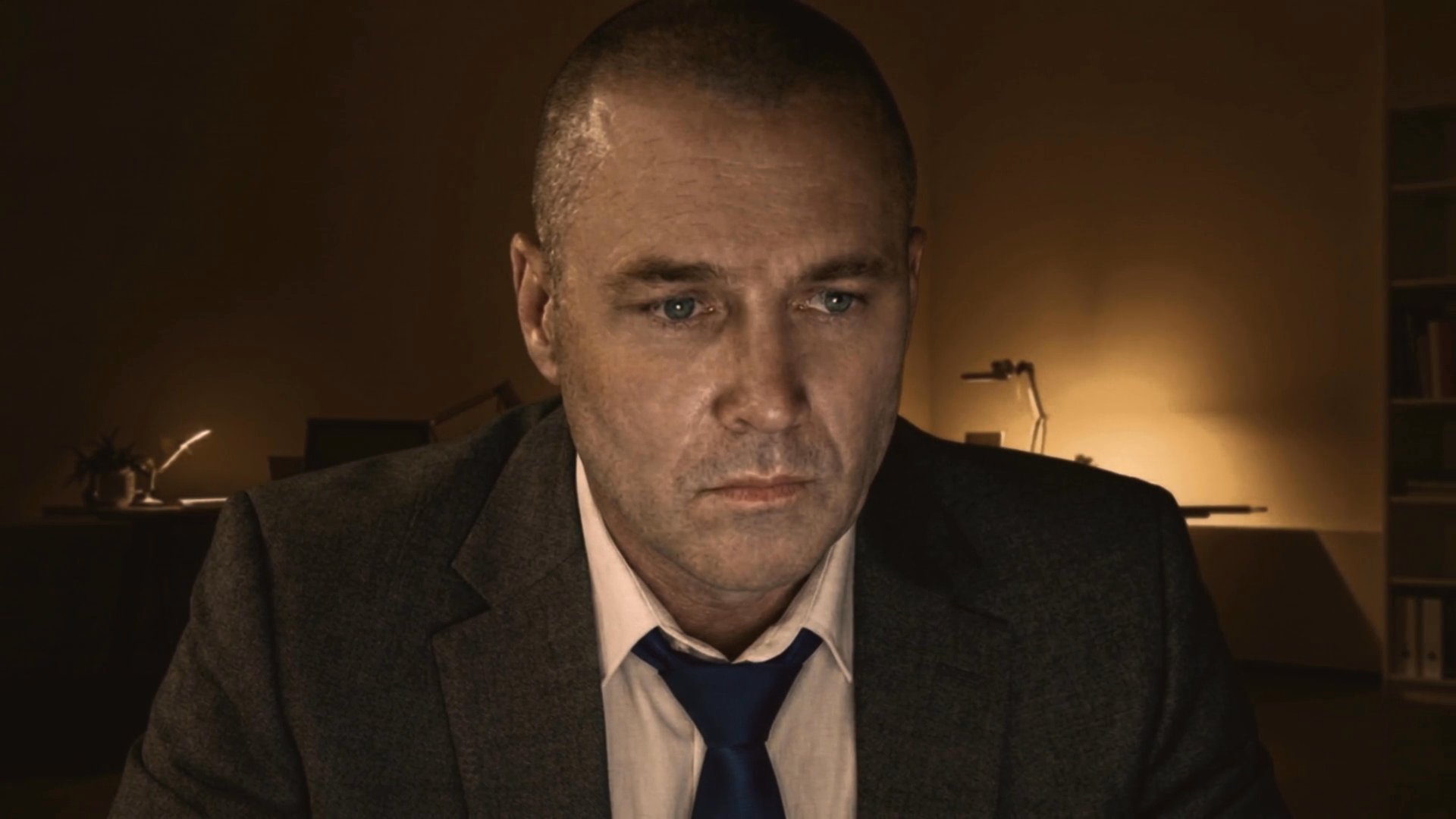 A man in a suit monitors desert landscape footage through a specialized investigative software interface. He observes a subject on screen while cross-referencing case file data.