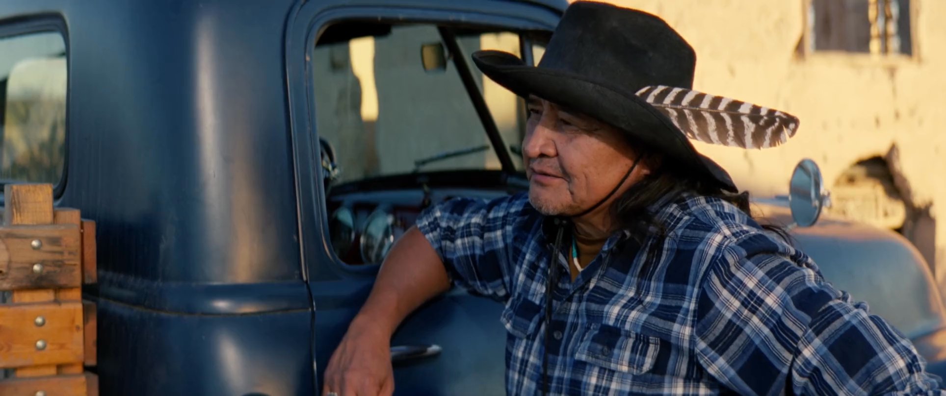 An indigenous man wearing a feathered hat leans against a vintage pickup truck while conversing with a man in an outdoor desert setting.