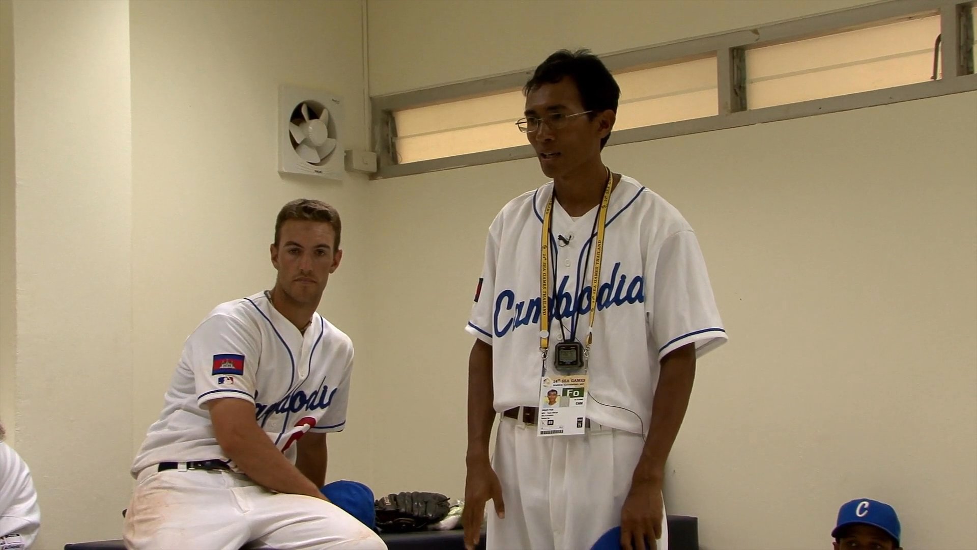 A coach motivates a team of Cambodian baseball players in a locker room, emphasizing perseverance and team spirit before their final game. The athletes reflect on their previous match and share their resolve to finish the tournament strong.