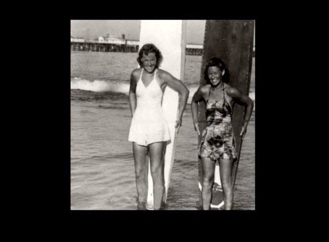 An elderly man recounts personal memories while archival photographs of women surfing in the early twentieth century are displayed on screen. The imagery transitions from nostalgic reflections to historical scenes of individuals engaging with the ocean and vintage surfboards.