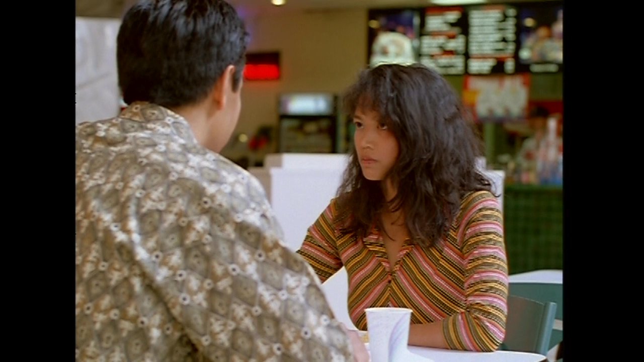 A man and woman engage in a serious, focused conversation while seated together at a table in a food court.