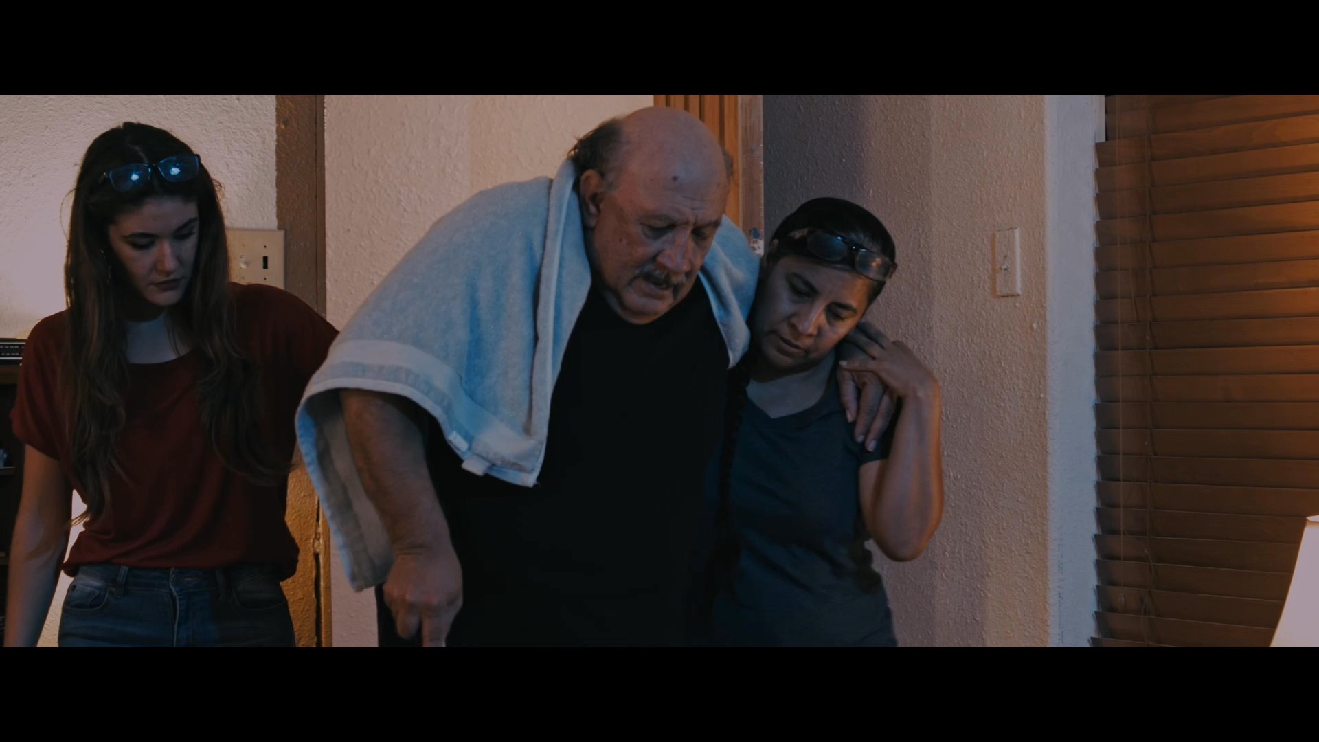 Two caregivers carefully support an elderly man as they guide him across a room into a bedroom. The individuals maintain a slow, steady pace to ensure his stability and comfort.