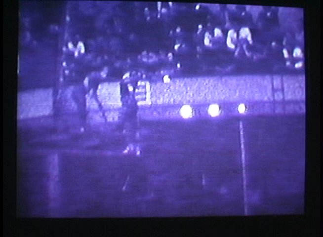 Athletes perform high-dives into a swimming pool during a nighttime competition captured with night-vision optics. Spectators observe from the bleachers as divers execute their routines.