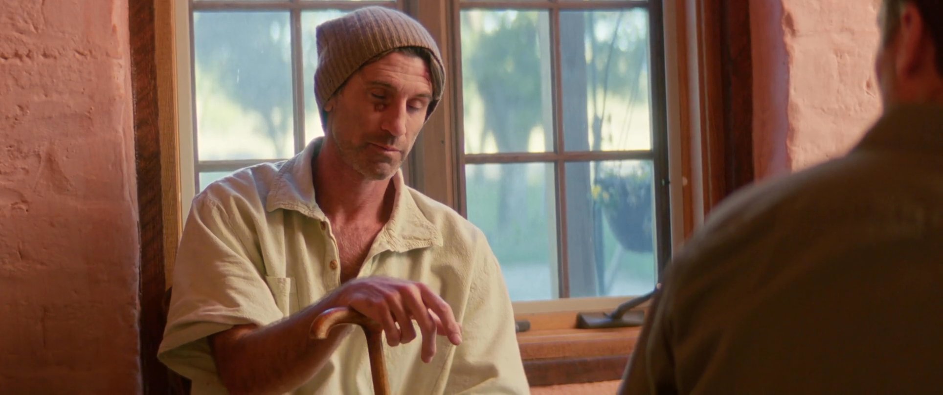 Two men engage in an intense, seated conversation inside a rustic room. One man sits by a window holding a cane, while the other leans in, listening intently.