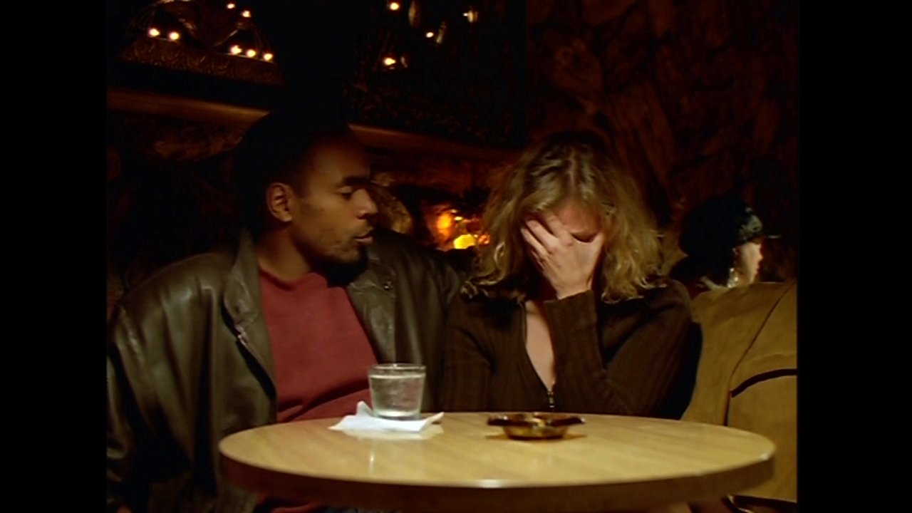 A man offers emotional comfort to a distressed woman as they sit together at a dimly lit bar booth. The man speaks gently and holds her hands, providing reassurance during a difficult moment.