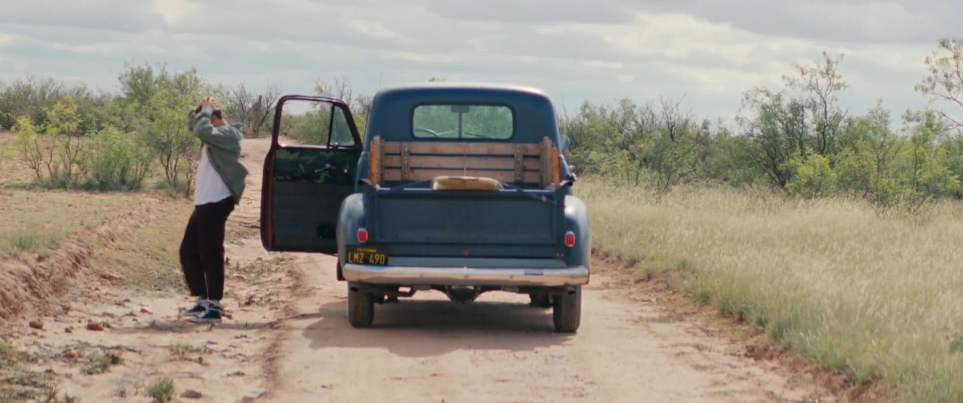 A man experiences a vehicle malfunction while traveling through a remote desert landscape. He exits his vintage pickup truck to assess the situation amidst the vast, arid terrain.