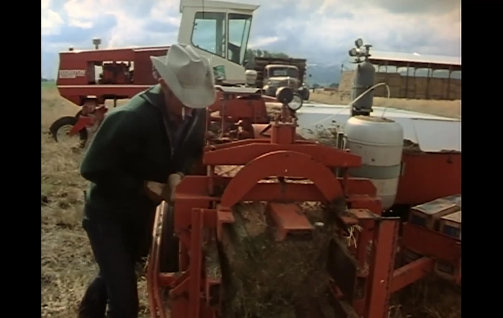 A rancher pulls leftover hay from a red baler by hand in an open farmyard. He clears the machine for storage and says he plans to saddle his horse and head to Nevada.