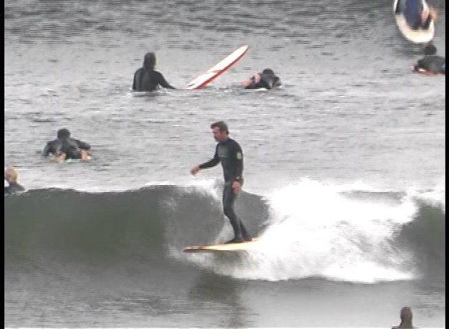 A surfer rides a wave in the open ocean while other surfers wait in the lineup. The surfer skillfully maneuvers the board across the face of the swell.