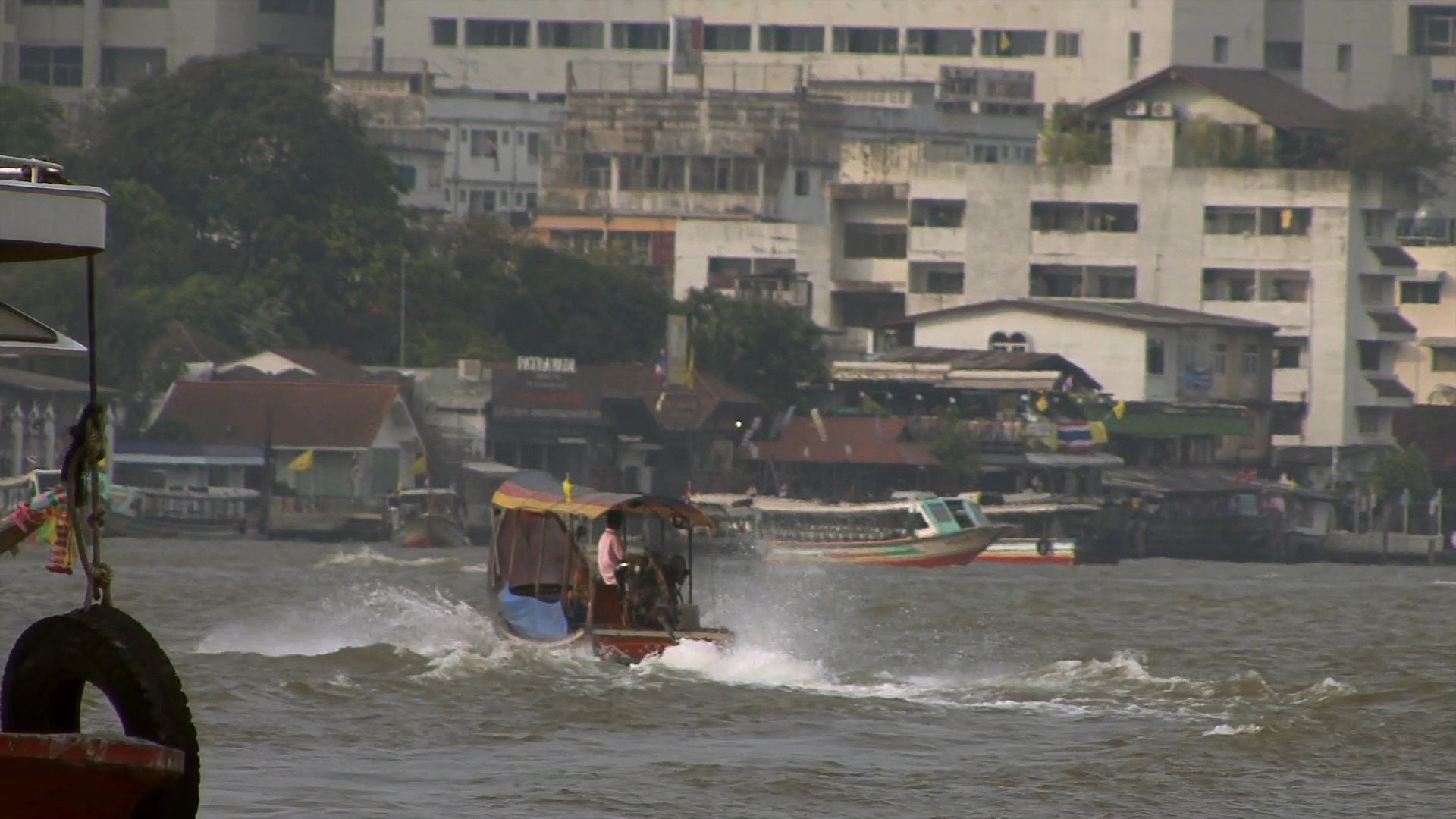 Commuter traffic flows through city streets alongside a tuk-tuk before transitioning to river transit on a ferry traveling across the Chao Phraya River.