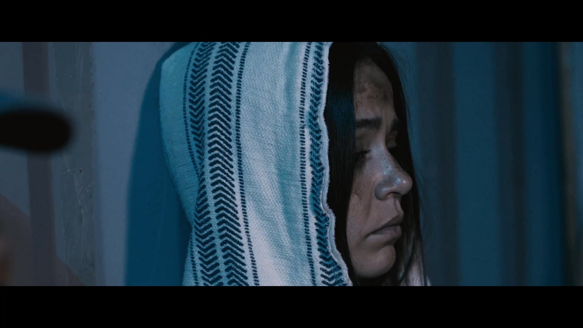 A man intensely questions a woman named Marisela in a dimly lit, confined space, creating a moment of palpable emotional tension. He challenges her silence and lack of defense as they sit together in an austere environment.