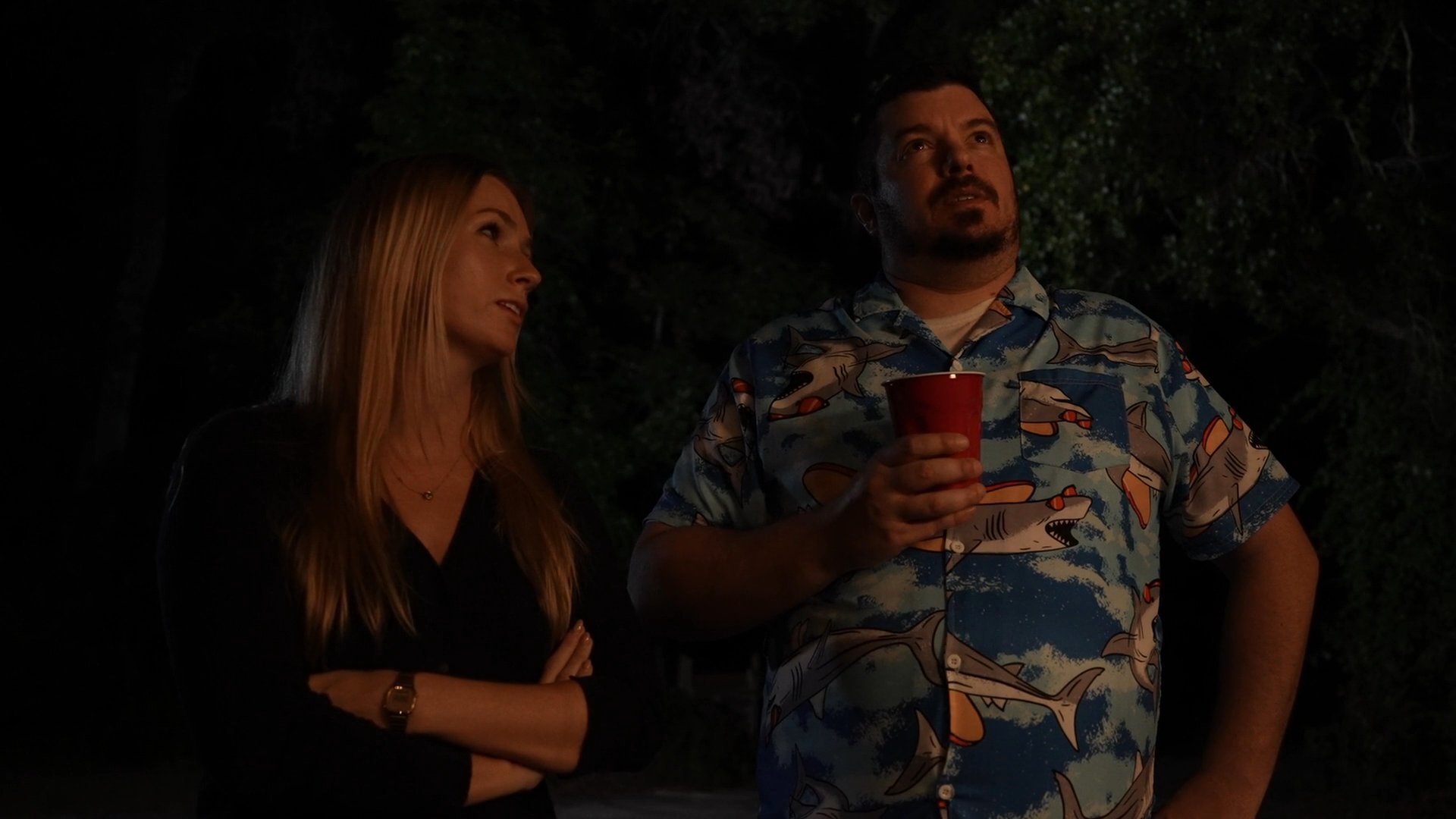 A man and a woman stand together outdoors at night, gazing upward at the sky during a casual social setting.