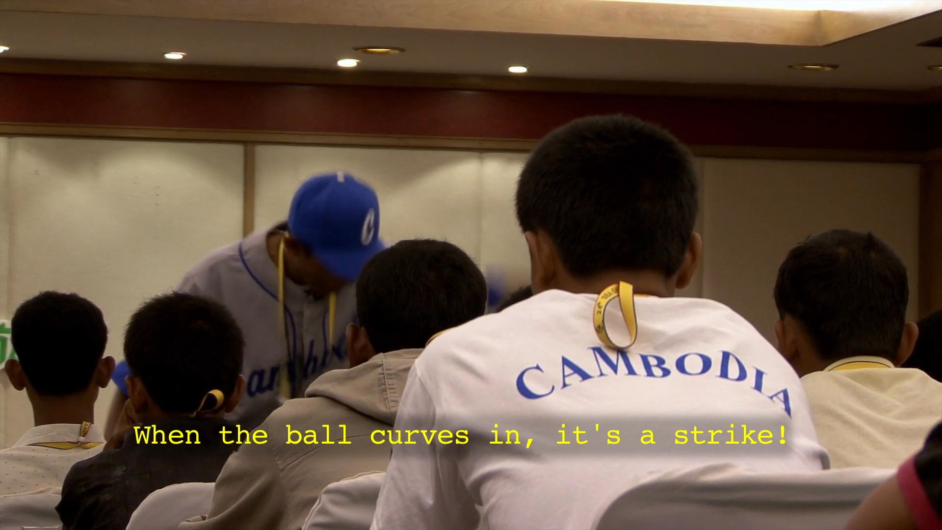 An experienced coach addresses a group of Cambodian baseball players during a team meeting, emphasizing technique and mental discipline.