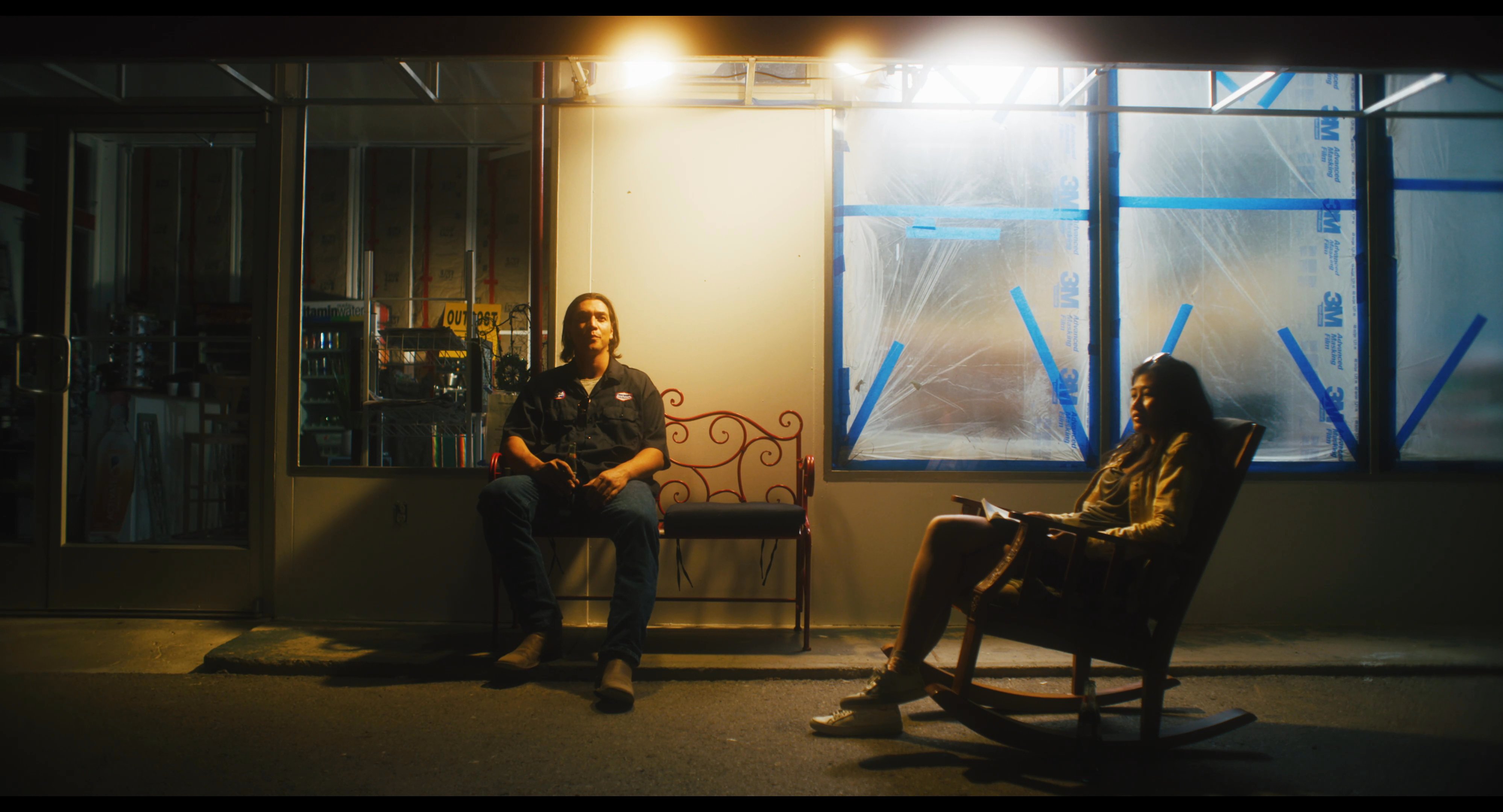 Two individuals sit outside in the evening, engaging in a quiet, candid conversation under artificial lighting.