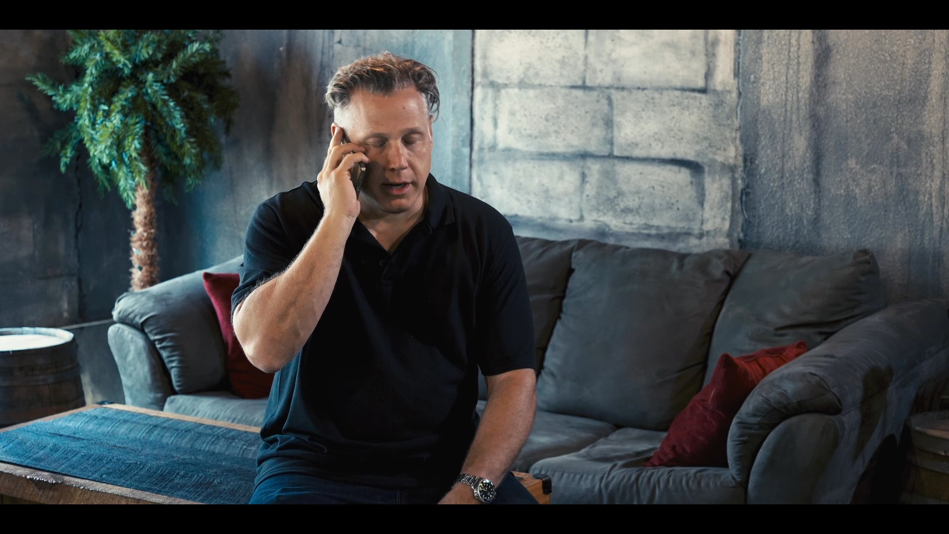 A man in a black polo shirt concludes a telephone conversation while seated on a sofa before standing and transitioning into an industrial-style environment.
