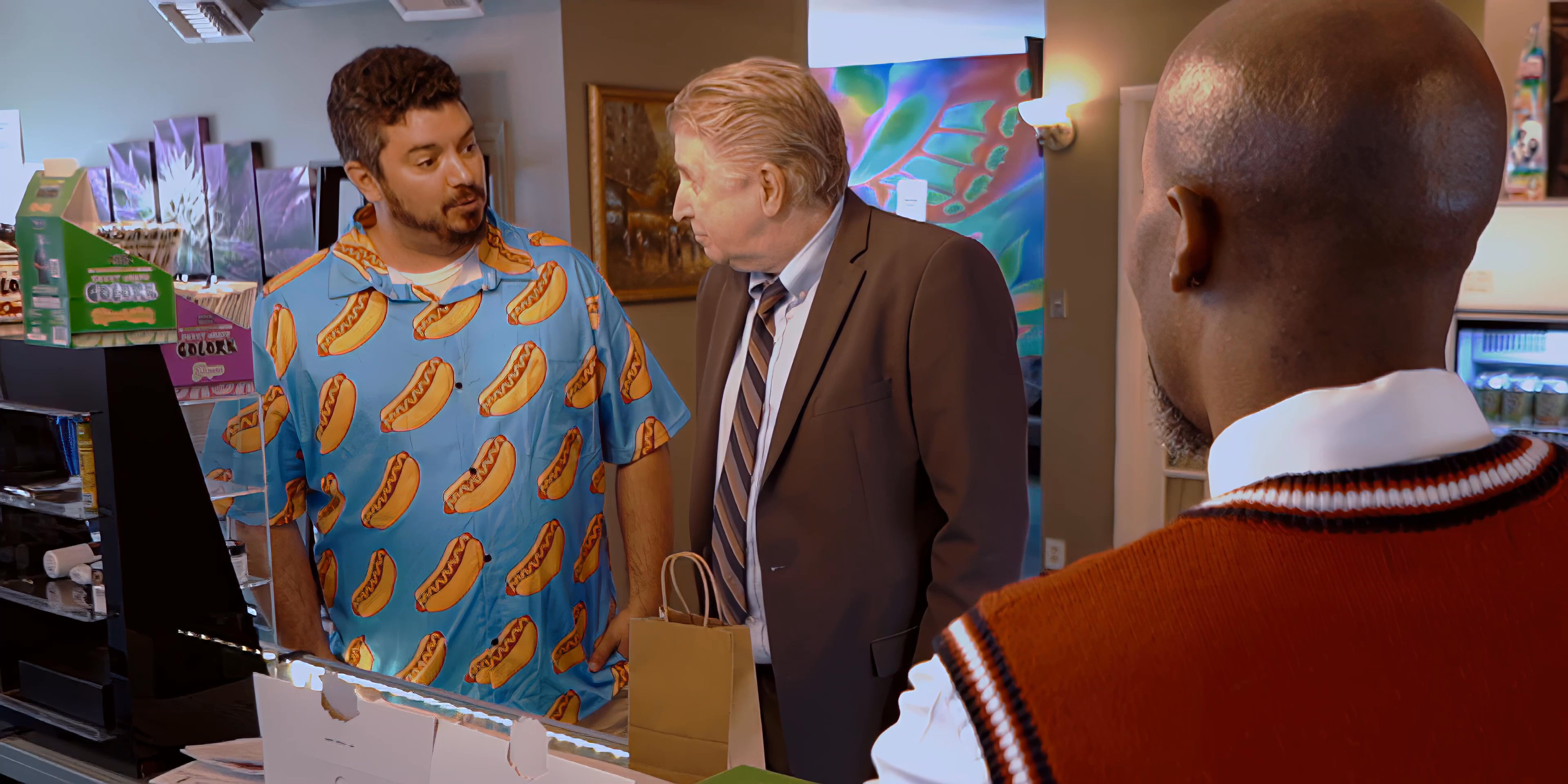 A man in a vest manages a retail counter while engaging in a conversation with two customers inside a store. The group discusses business matters amidst the product-filled commercial environment.